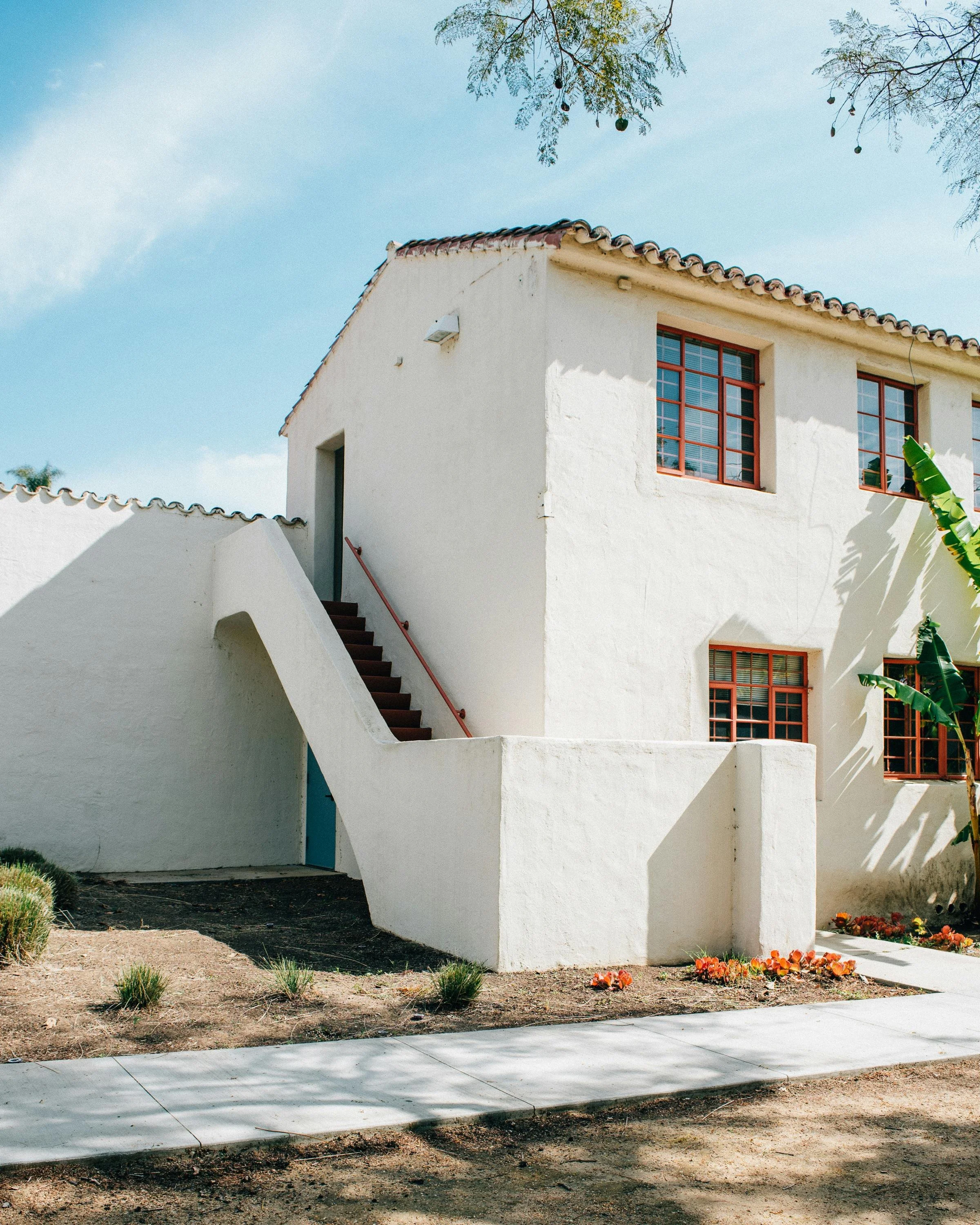 White stucco building with red window frames, outdoor staircase with red handrail, and small garden with orange flowers in front.