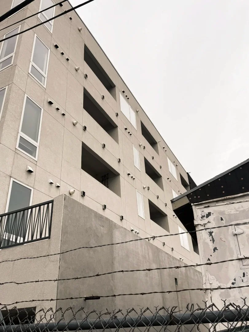 A multi-story beige apartment building with multiple windows and balconies, viewed from below, with barbed wire and a fence in the foreground.