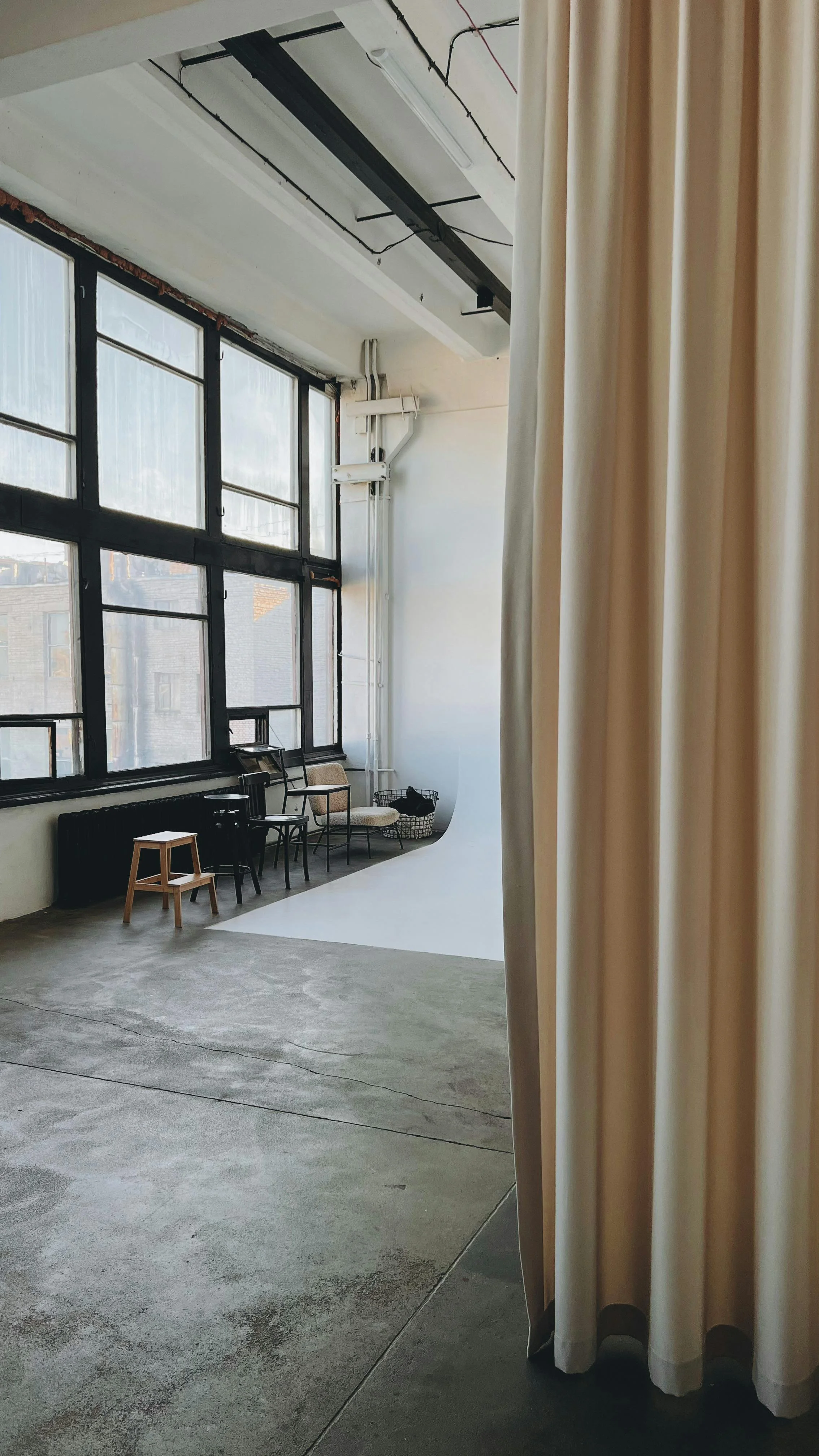 Indoor studio space with large windows, chairs, a small stool, and a white backdrop, partially obscured by a beige curtain.