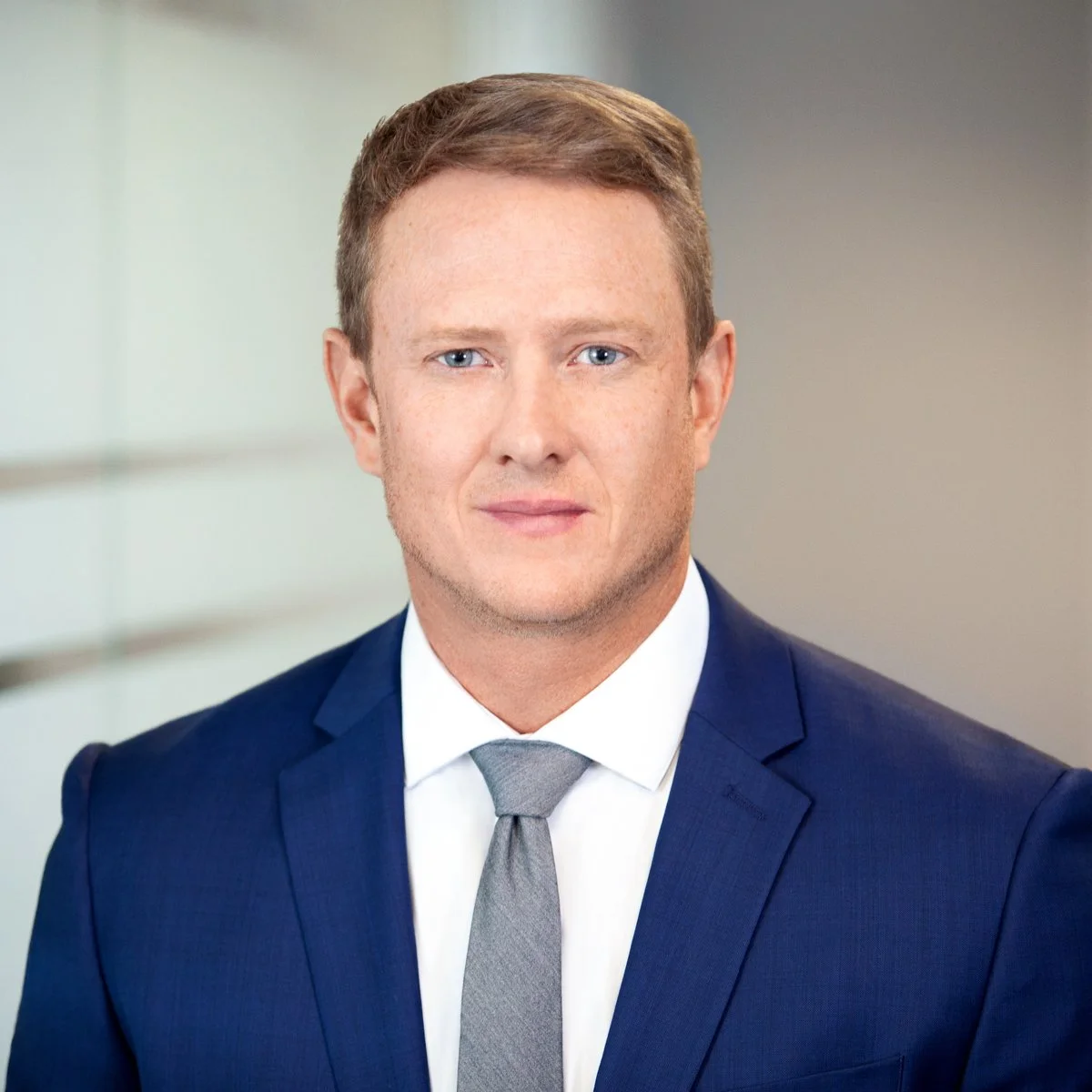 A professional man in a blue suit and gray tie posing in an office setting with neutral background.