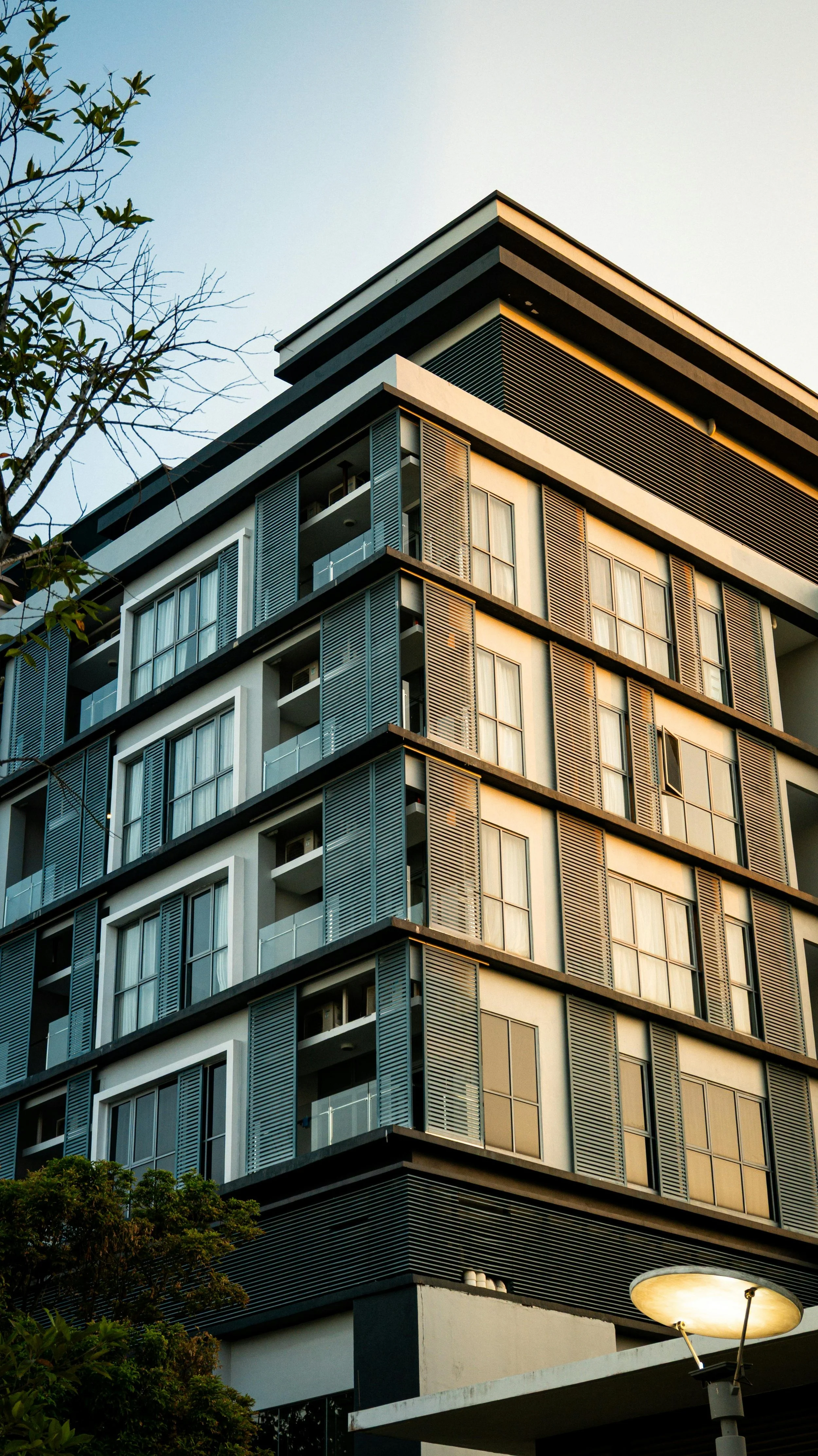A modern multi-story apartment building with large windows and open balconies, partly lit by the setting sun, with trees and a streetlamp in the foreground.