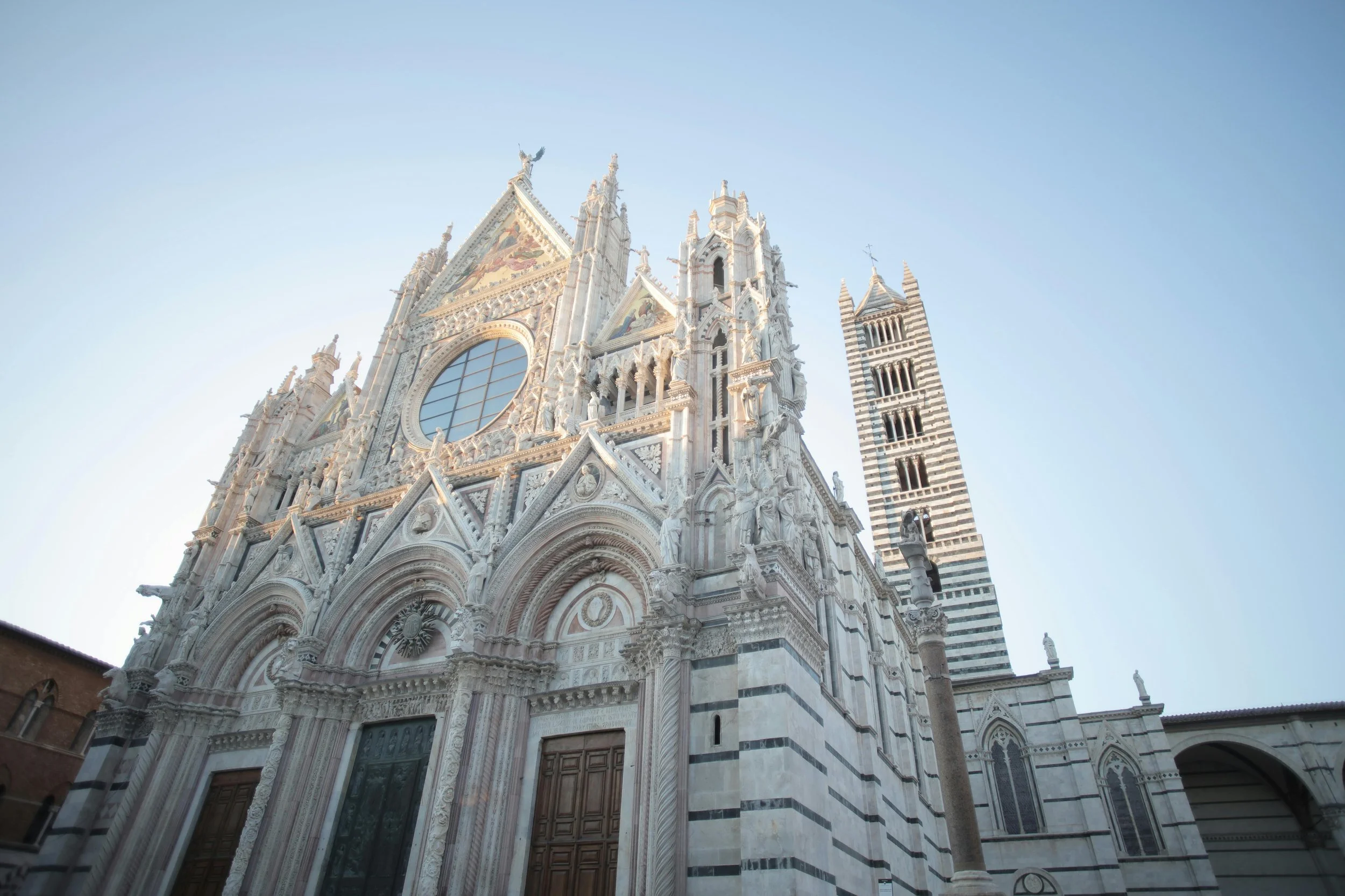 Front view of a large, ornate Gothic cathedral with intricate white marble façade, tall spires, statues, and large stained glass window, set against a clear blue sky.