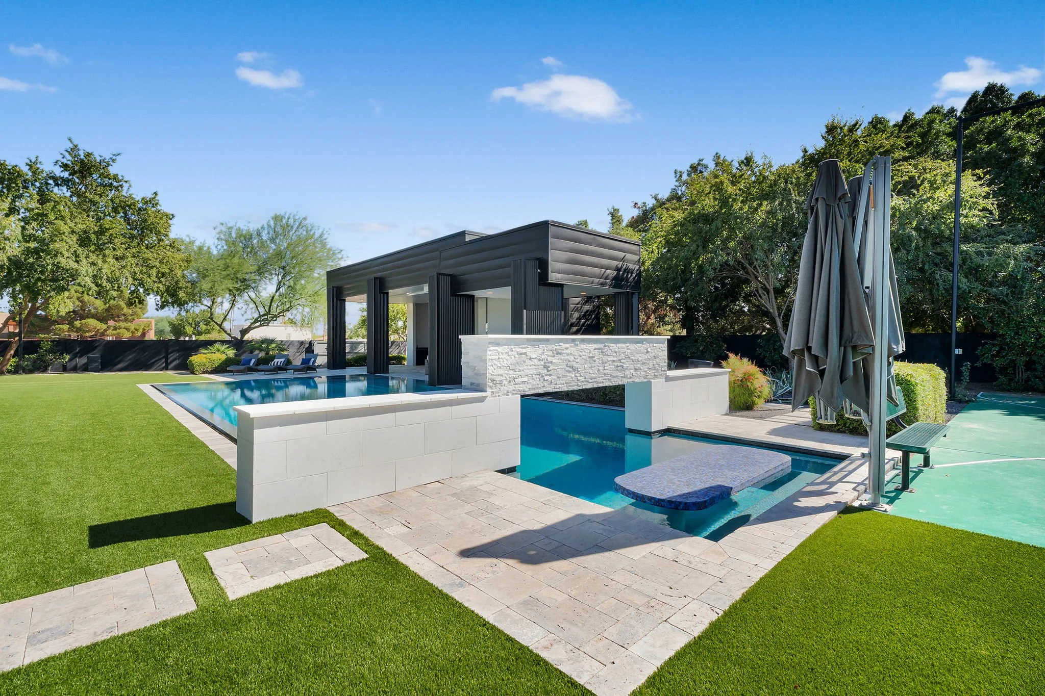 Modern backyard with a swimming pool, covered outdoor seating area, and a tennis court surrounded by green trees and lawn.