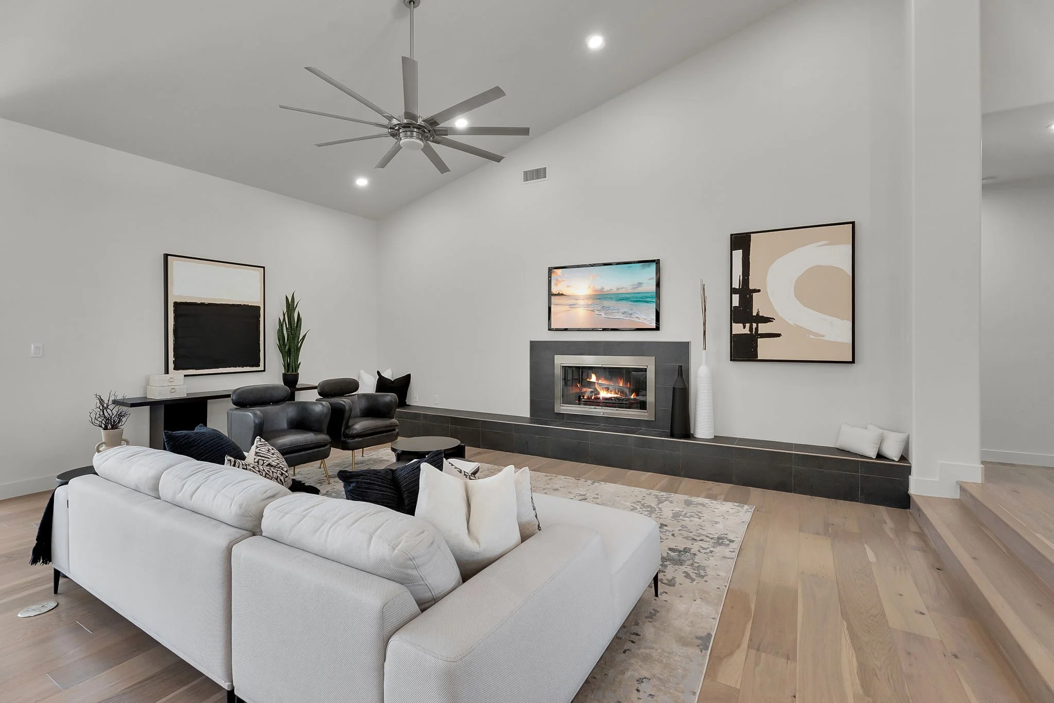 Modern living room with white walls, hardwood floors, a gray fireplace, flat-screen TV, black armchairs, a white sofa with black and white pillows, abstract artwork, decorative vases, a ceiling fan, and a large window.