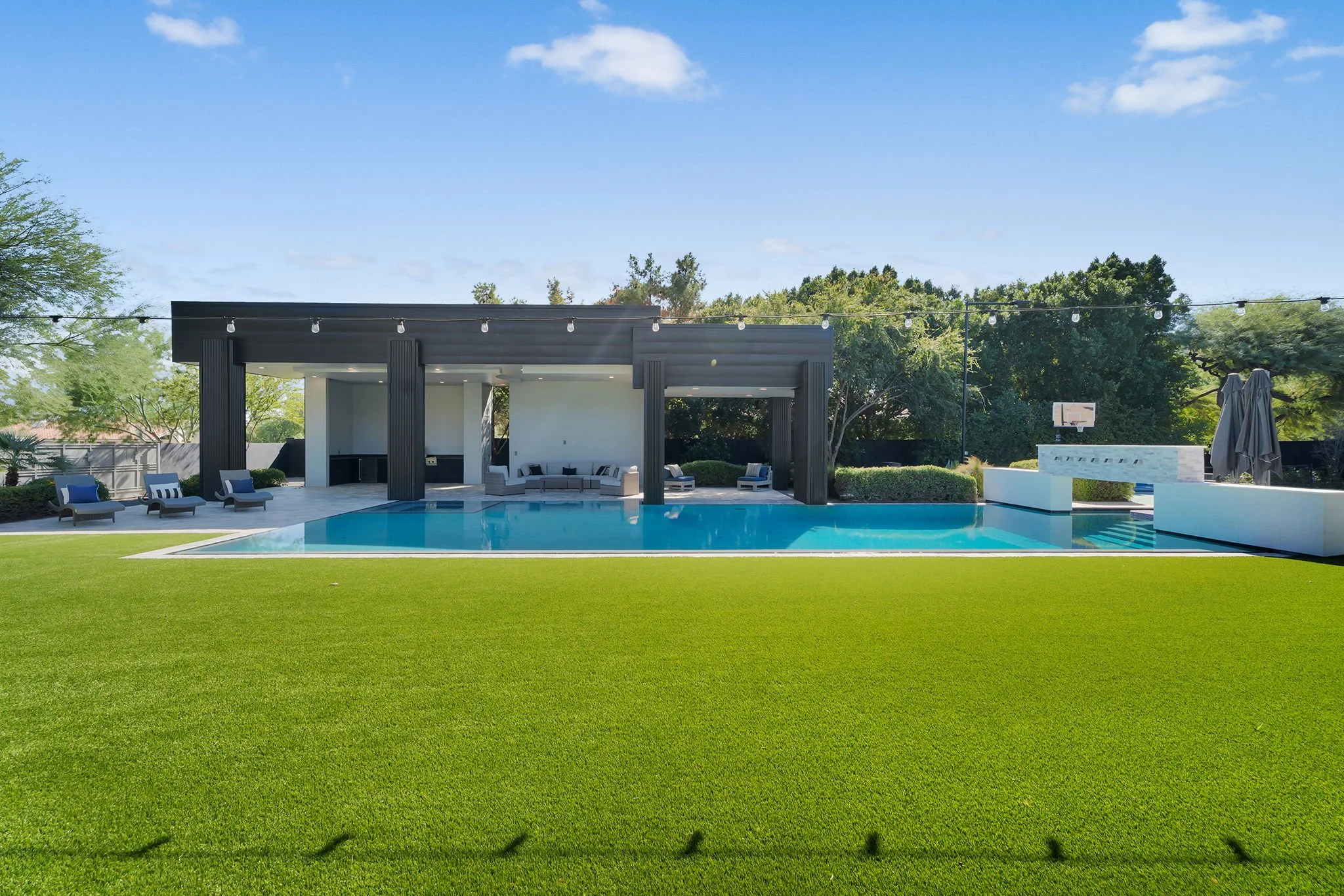 Modern backyard with a swimming pool, outdoor seating area, and a shaded lounge space, surrounded by well-manicured green grass and trees.