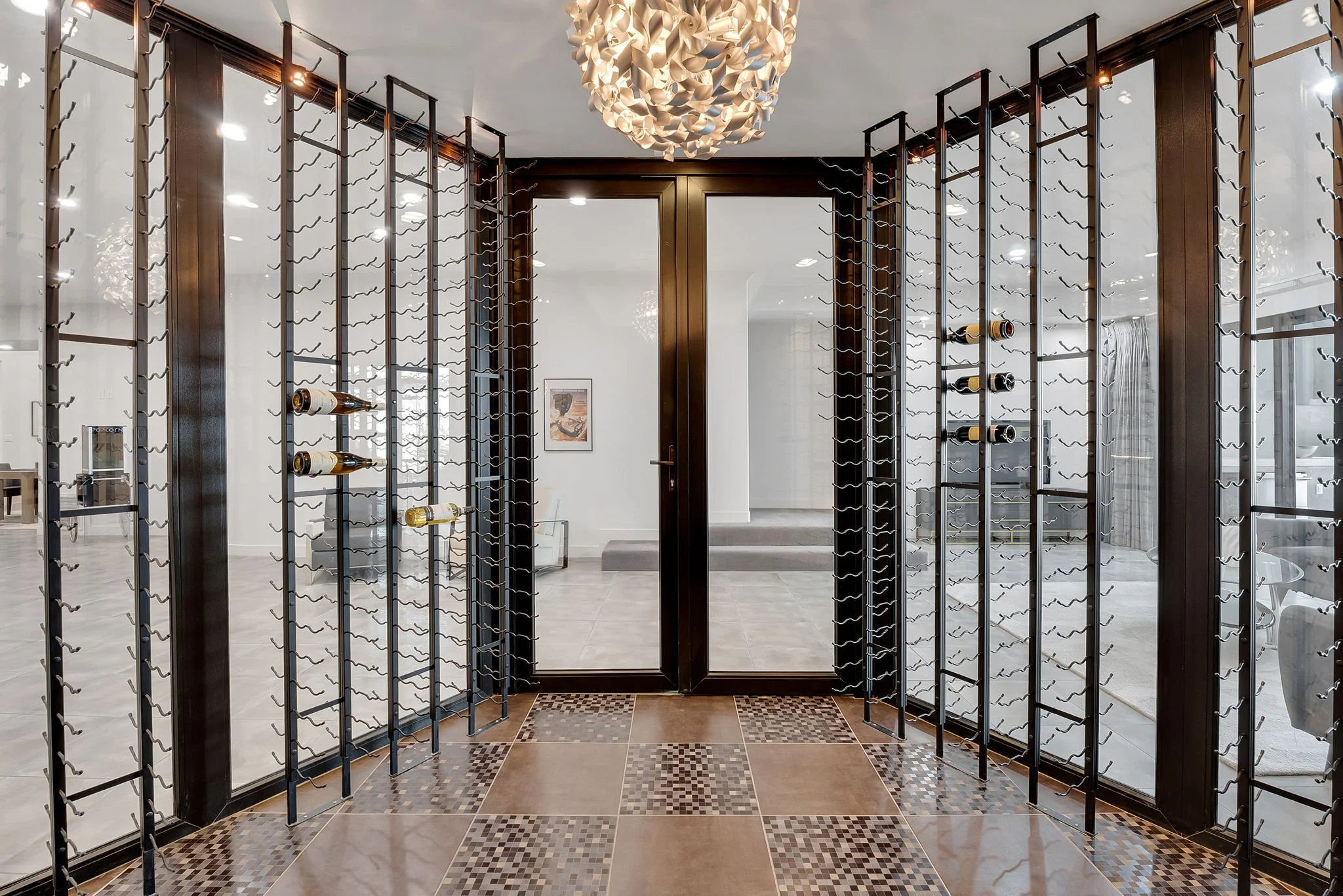 A modern wine cellar with a central glass door, surrounded by wine racks. The racks are made of metal and hold bottles horizontally. The floor features a tiled pattern, and there is a decorative light fixture hanging from the ceiling.