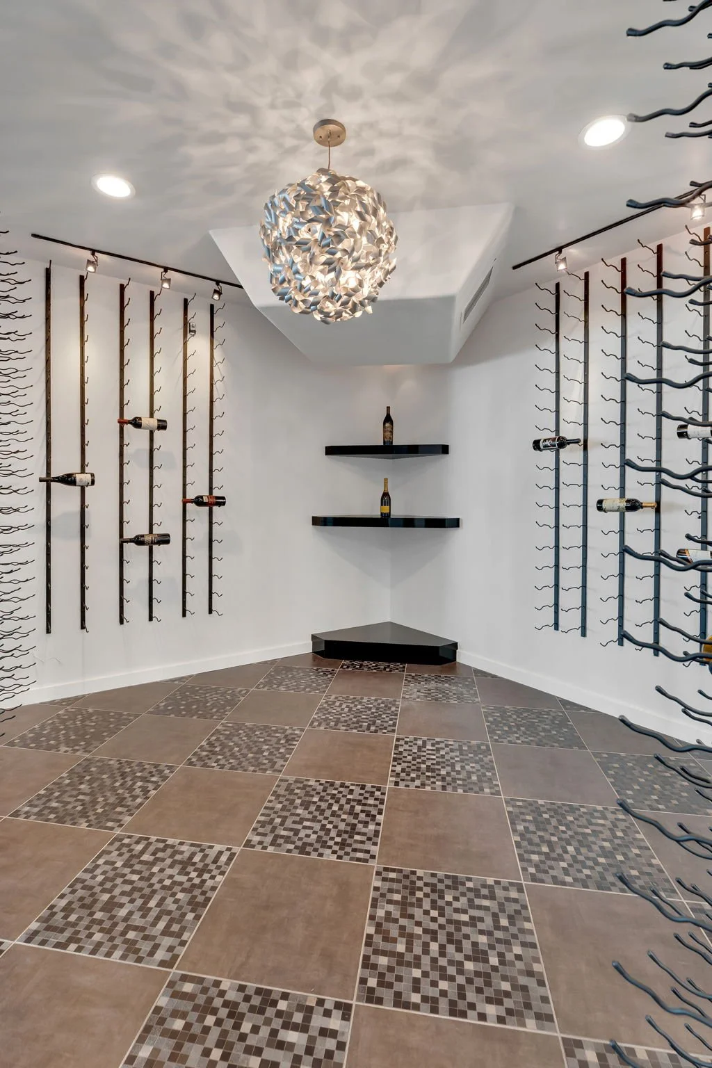A wine cellar with white walls and decorative tile flooring. Wine bottles are displayed on wall-mounted racks and floating shelves, with a modern pendant light hanging from the ceiling.