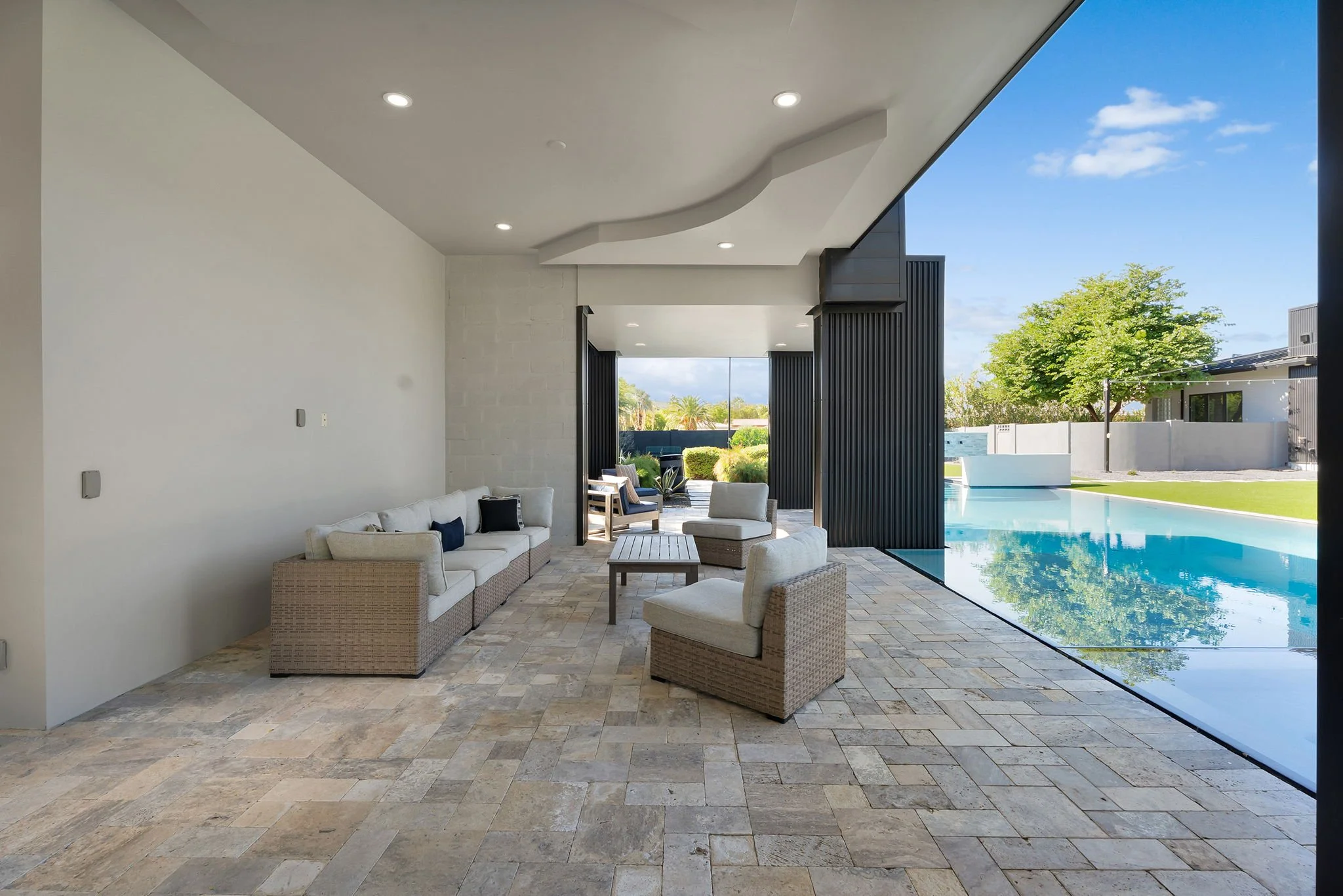 Modern outdoor patio with seating area, overlooking a swimming pool and backyard with trees and fence.