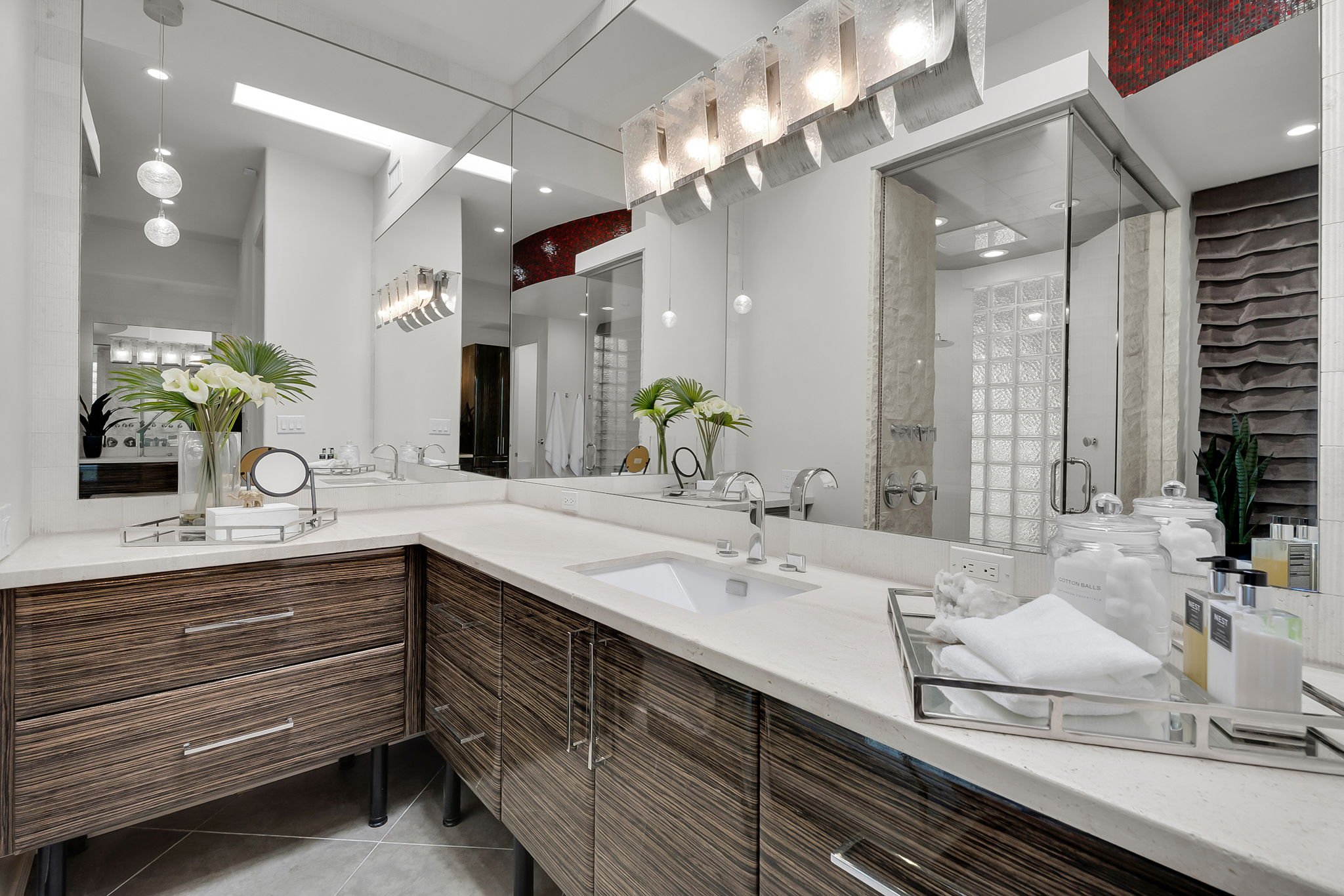 Modern bathroom vanity with a large mirror, white countertop, and wood grain cabinets. Decor includes a vase with white lilies, toiletries, and a tray of cotton balls and towels. The bathroom features minimalistic design with neutral tones and contem