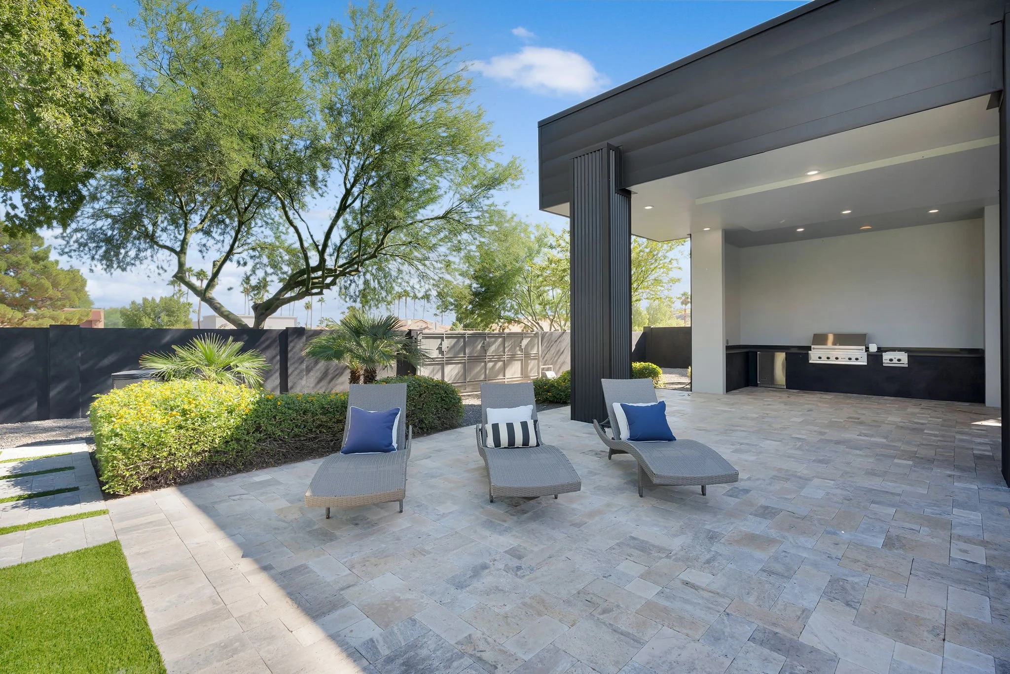 Outdoor patio with three lounge chairs with cushions, greenery, trees, and a built-in outdoor kitchen area under a covered structure.