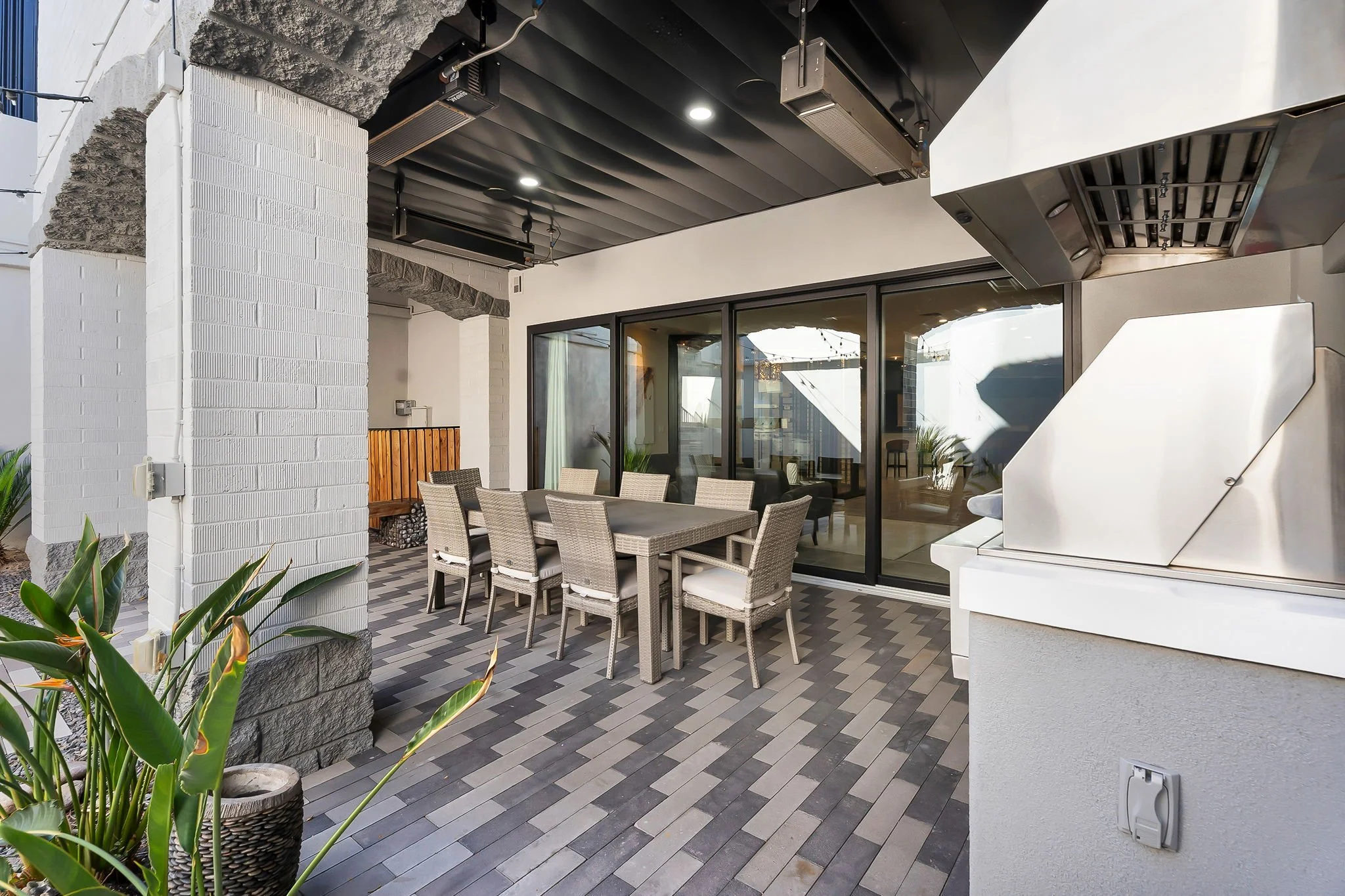 Outdoor dining area with a rectangular table and eight wicker chairs with white cushions, on a tiled floor, next to a built-in grill, with glass sliding doors leading inside, and potted plants nearby.