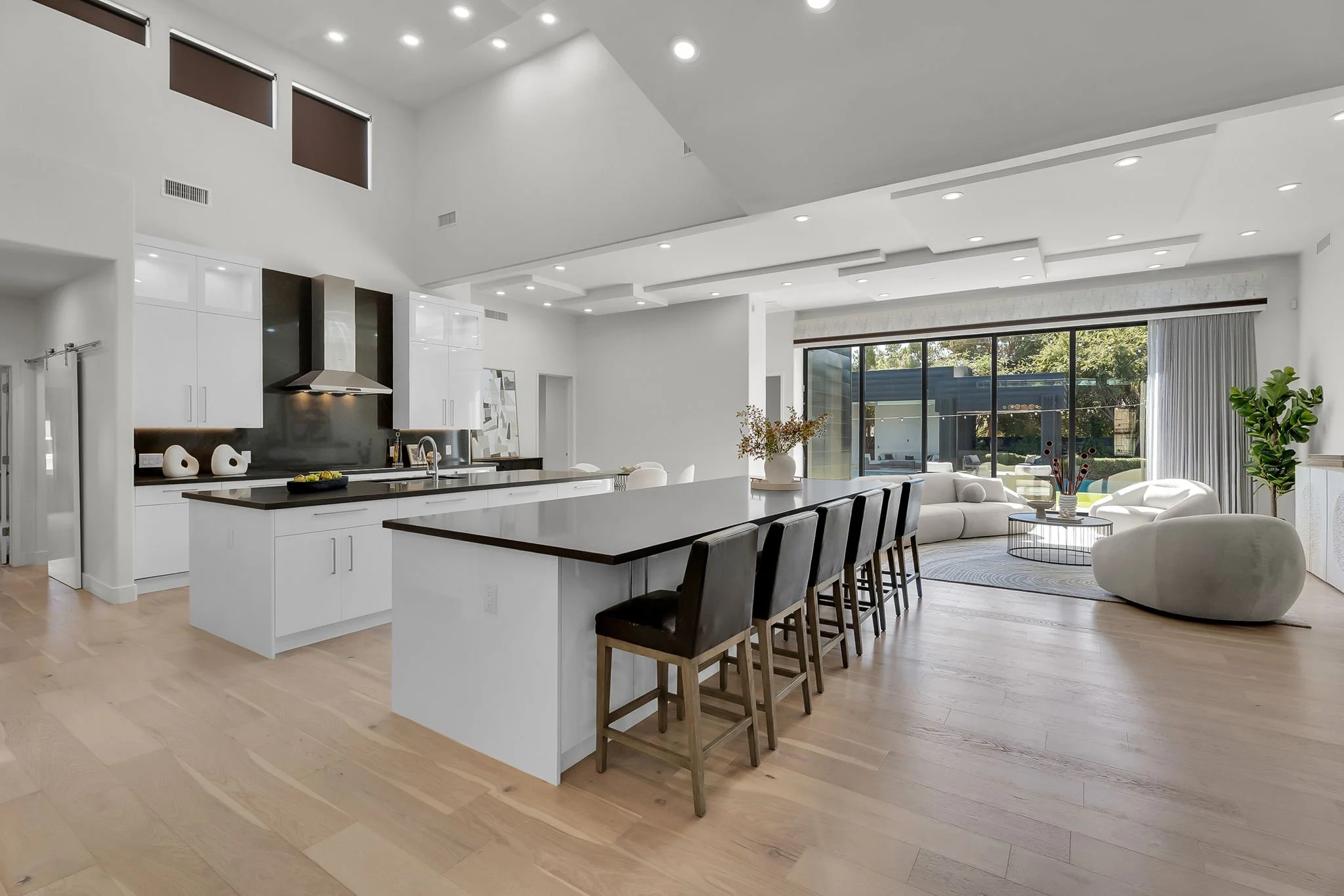Spacious, modern open-concept kitchen and living area with white cabinetry, black countertops, barstools at kitchen island, white sofas, large sliding glass door, hardwood floors, and minimalistic decor.