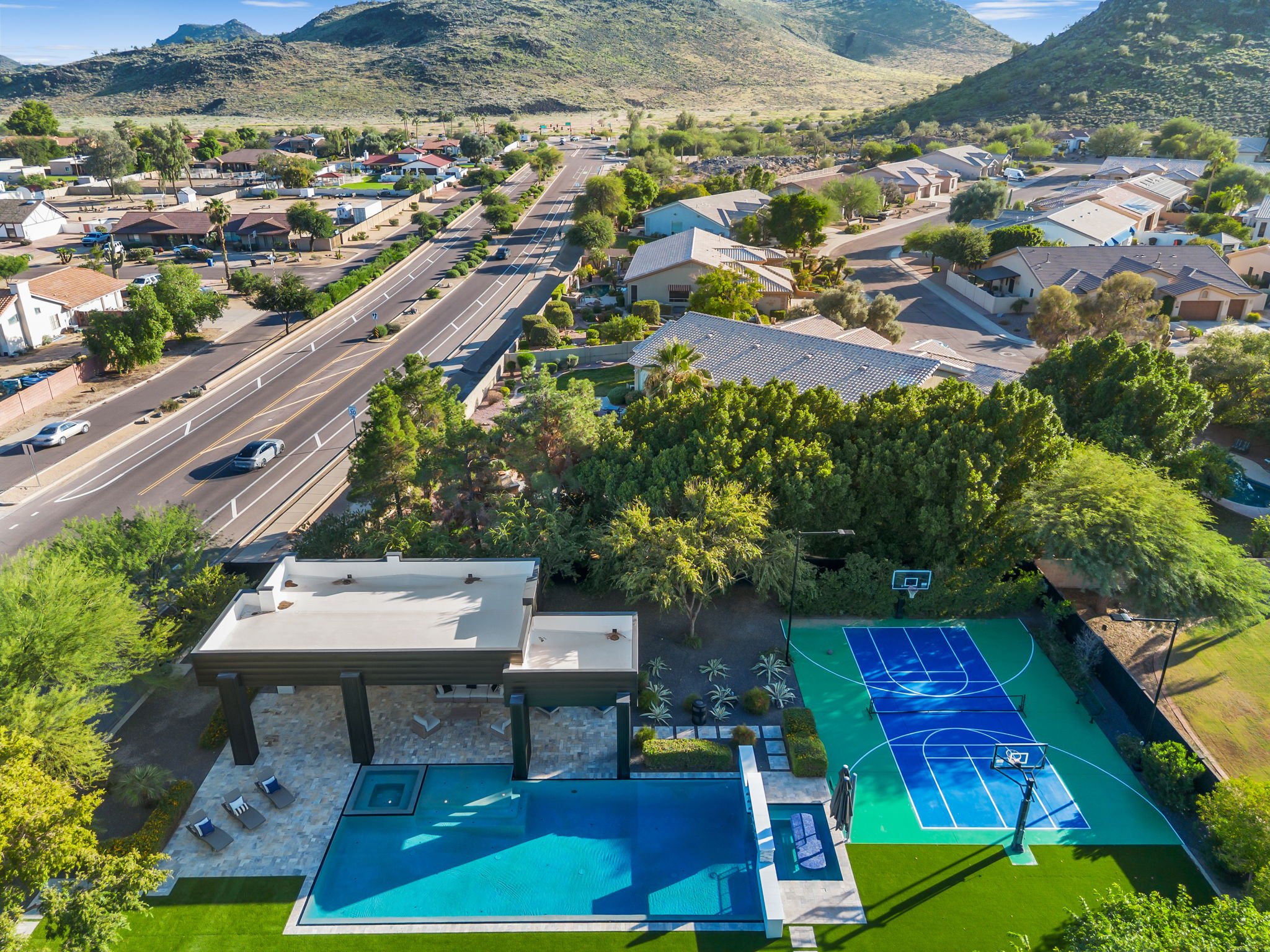 Aerial view of a residential neighborhood with houses, green trees, a sports court, and swimming pools, with mountains in the background.