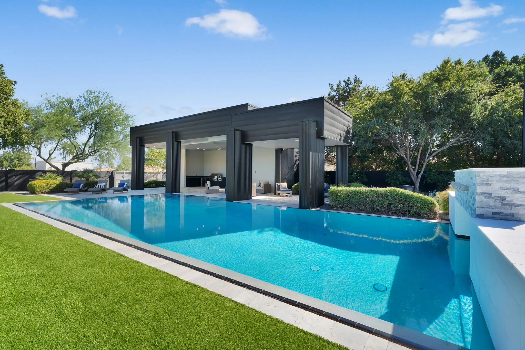 Modern backyard with a rectangular swimming pool, poolside lounge chairs, a covered sitting area with tables and chairs, lush green lawn, and trees in the background under a partly cloudy sky.
