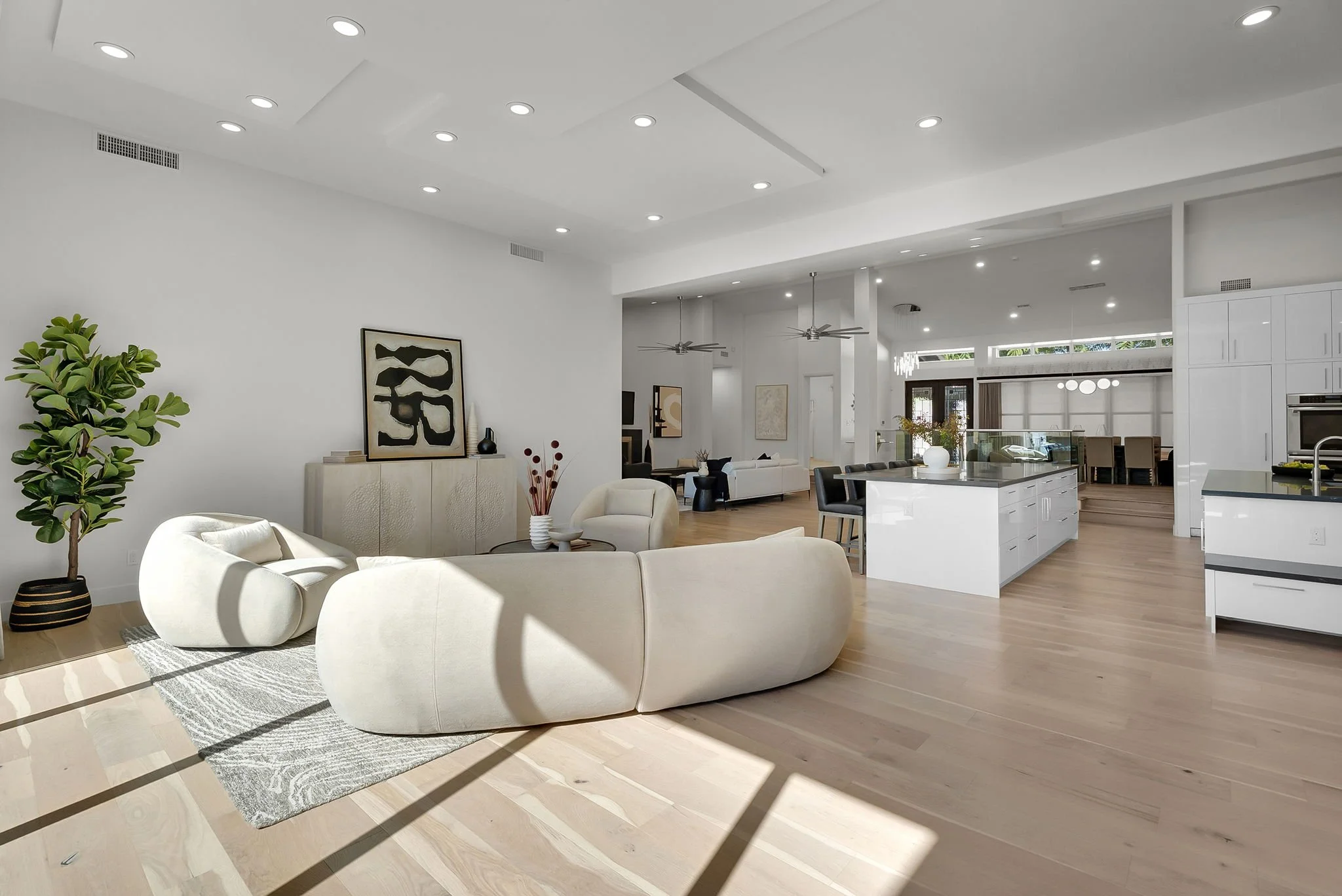 Bright, spacious living area with light wooden flooring, modern white furniture, a green potted plant, contemporary artwork, and an open kitchen with an island and bar stools.