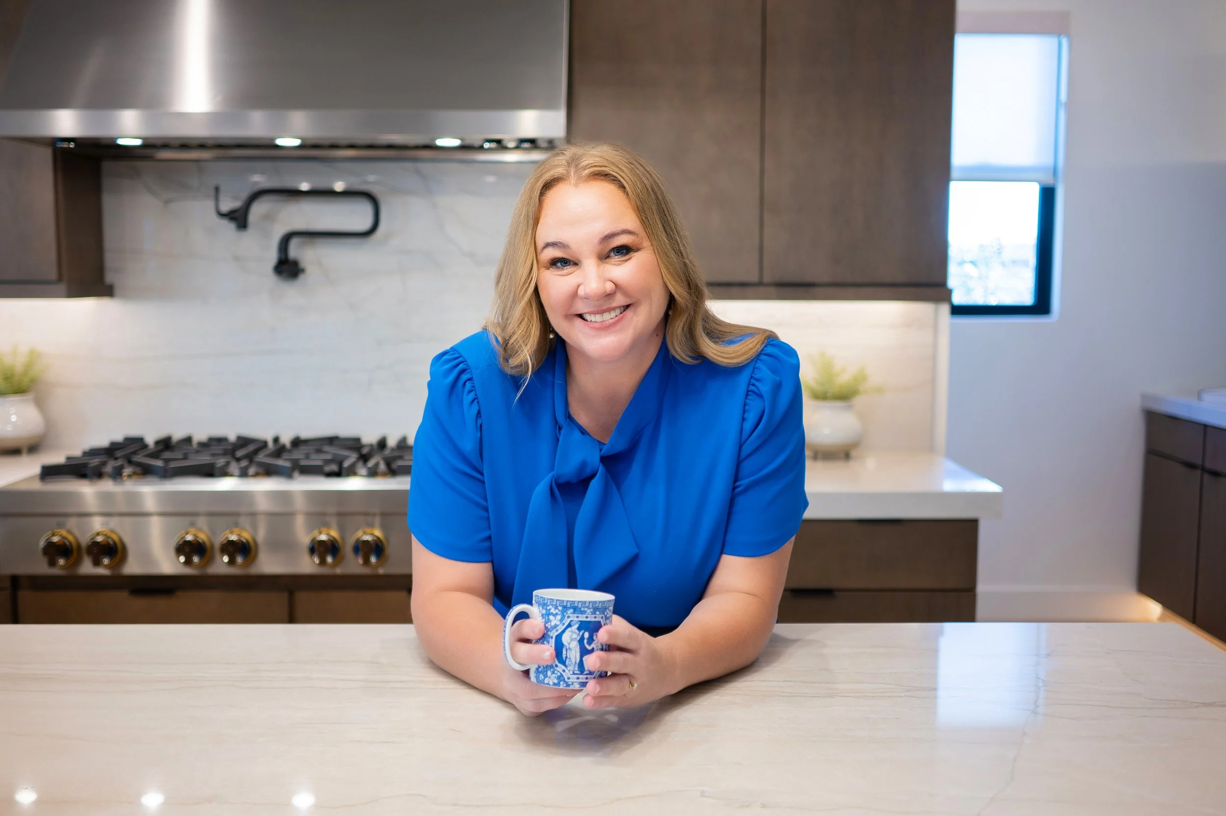 A woman with blonde hair wearing a blue blouse holding a blue and white mug in a modern kitchen.