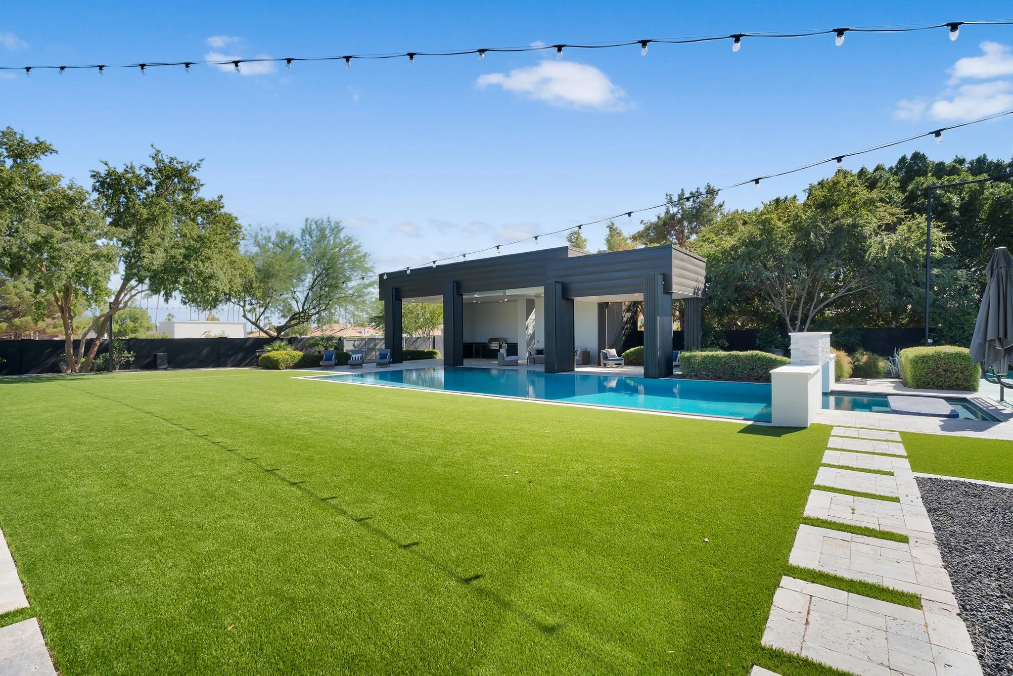 Modern backyard with a green lawn, a rectangular pool, a covered seating area with lounge chairs, and string lights overhead. Trees and bushes surround the area under a partly cloudy sky.