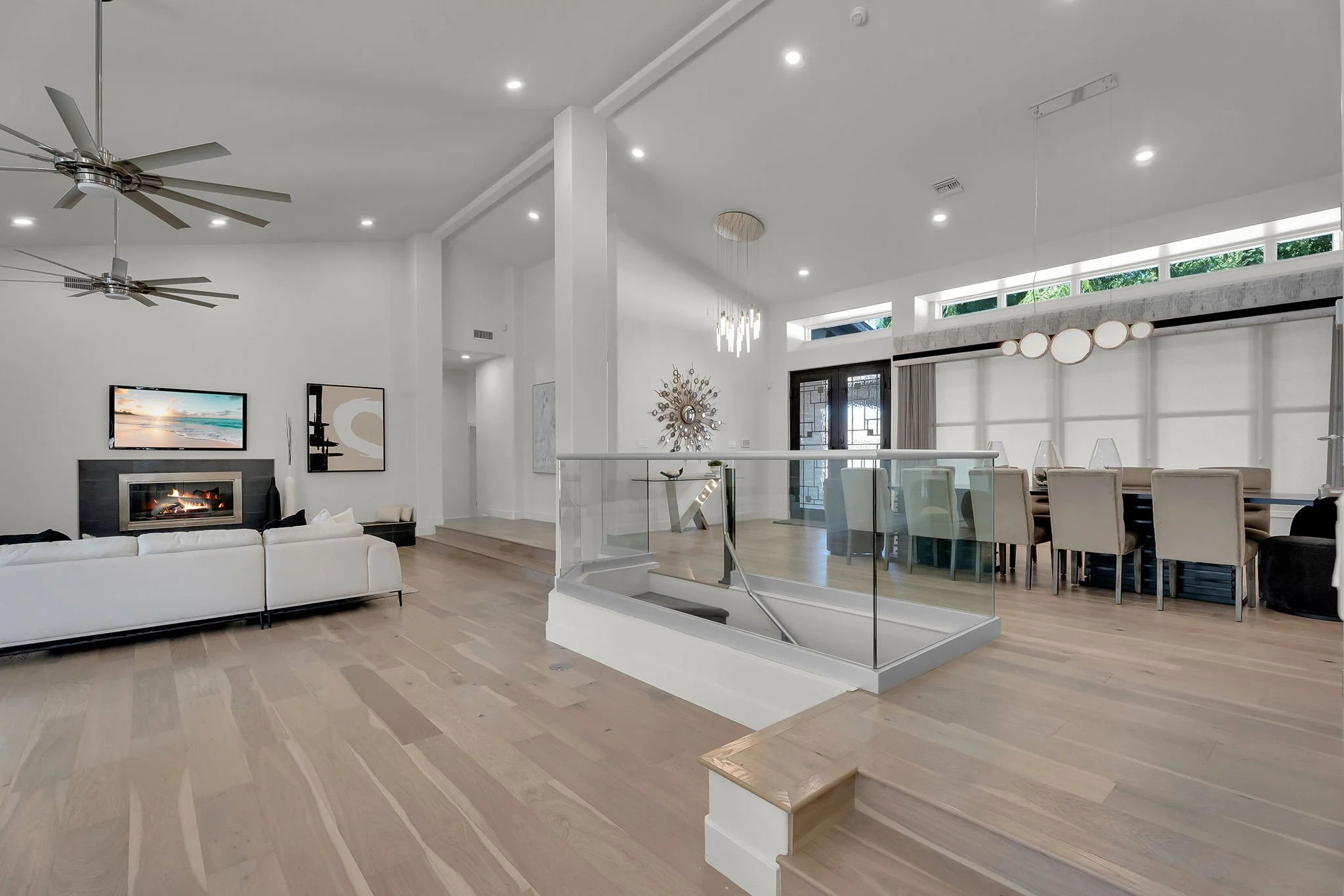 Modern living room and dining area with white walls, hardwood floors, glass staircase, ceiling fans, and minimal decor.