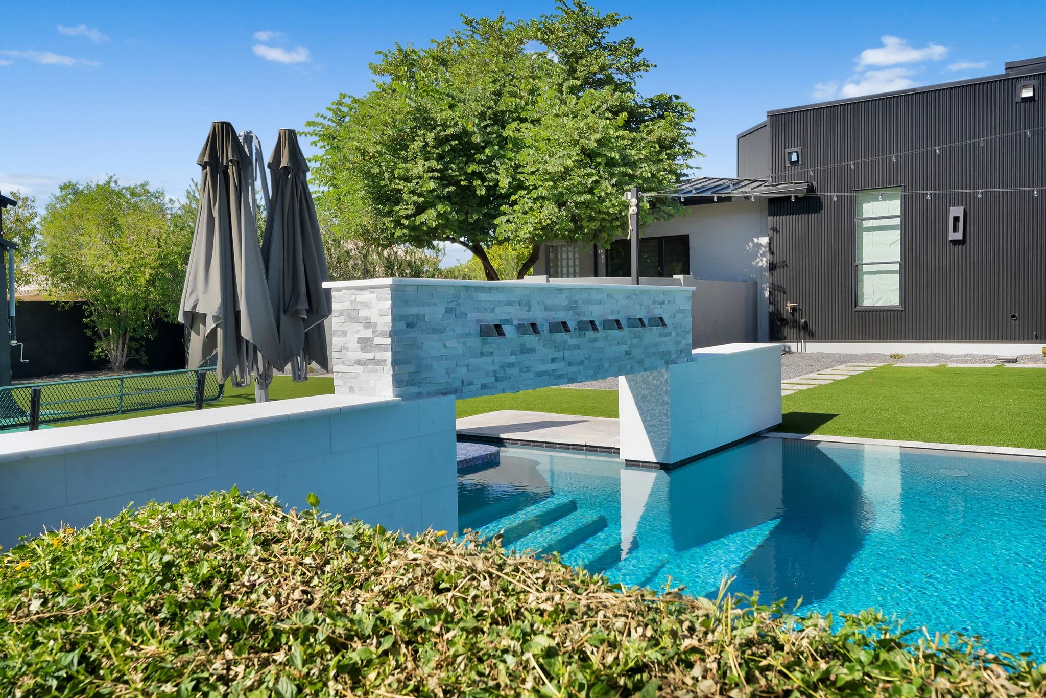 A backyard with a swimming pool, a modern waterfall feature, green grass, trees, and a black modern house under a blue sky.
