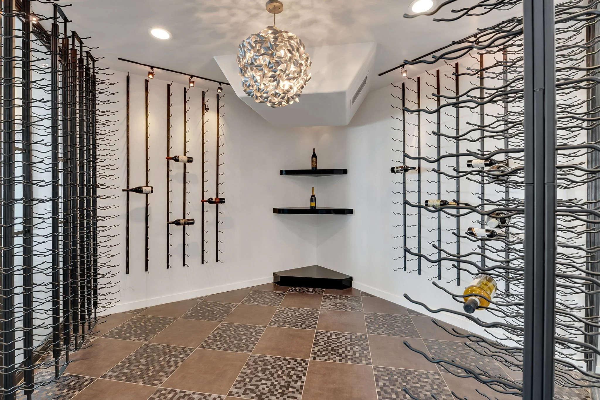 A modern wine cellar with walls decorated with black metal wine racks holding bottles, a black corner shelf with bottles, and a tiled floor with patterned square tiles. There's a unique white pendant light hanging from the ceiling.