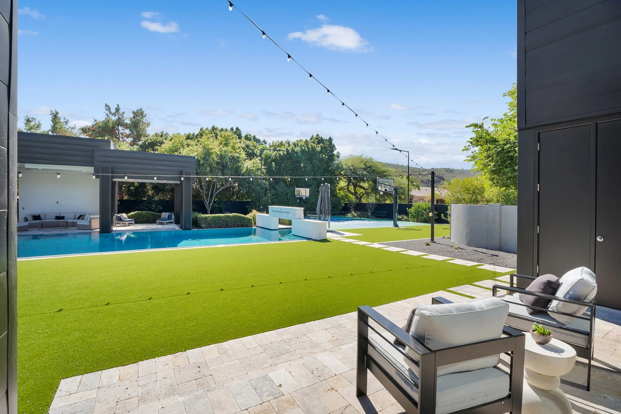Modern backyard with a swimming pool, outdoor seating, artificial grass, outdoor basketball hoop, string lights, and surrounding trees under a clear blue sky.