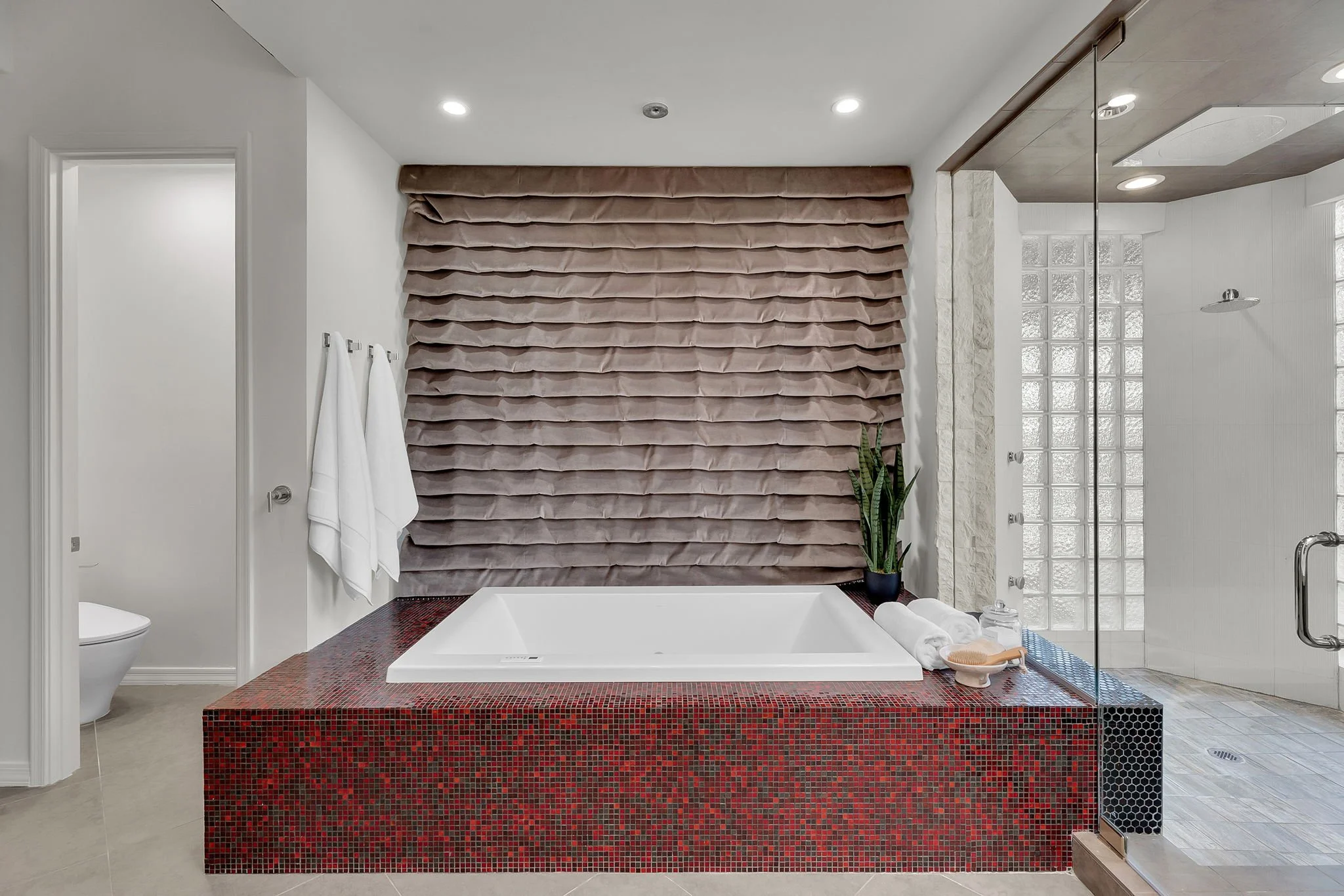 Modern bathroom with a large built-in bathtub surrounded by a red mosaic tile platform, beige tiled floor, a textured brown window covering, a plant, and a clear glass shower enclosure.