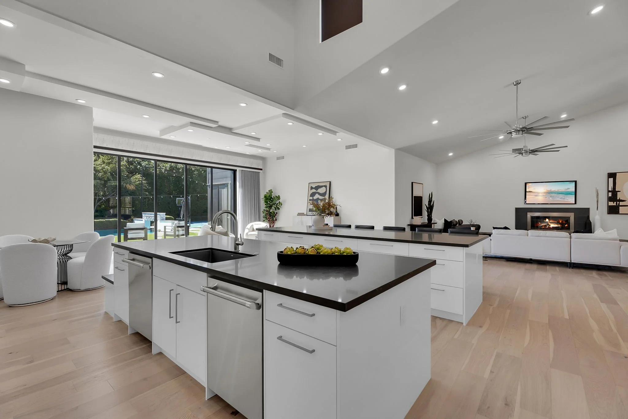Open concept kitchen and living room with white cabinetry, kitchen island, and modern furniture. Large sliding glass doors lead outside to a backyard with a pool and patio, and a fireplace with a TV above in the living area.
