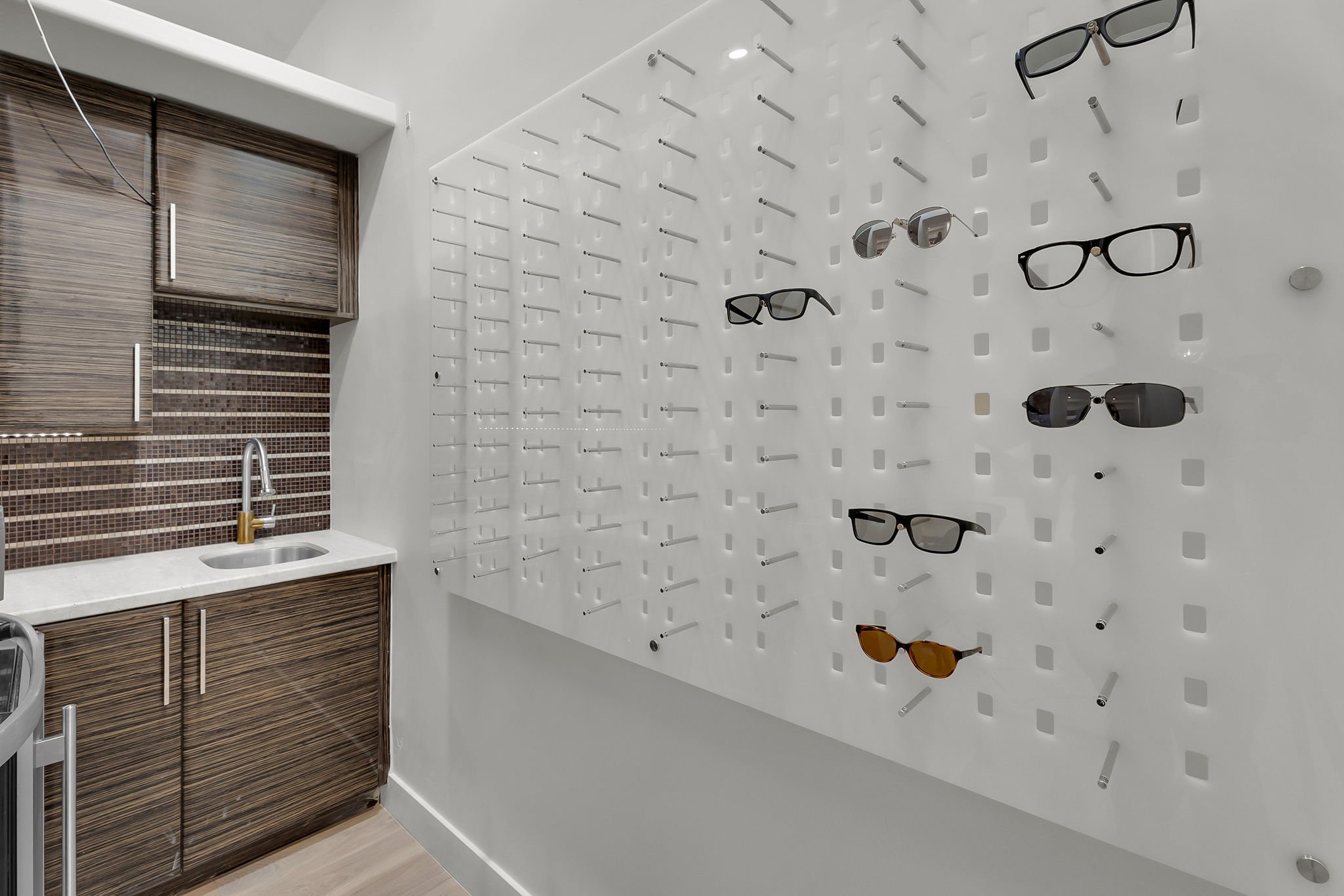 Eyewear display wall with various sunglasses in an retail store or optician's office, next to a kitchenette area.