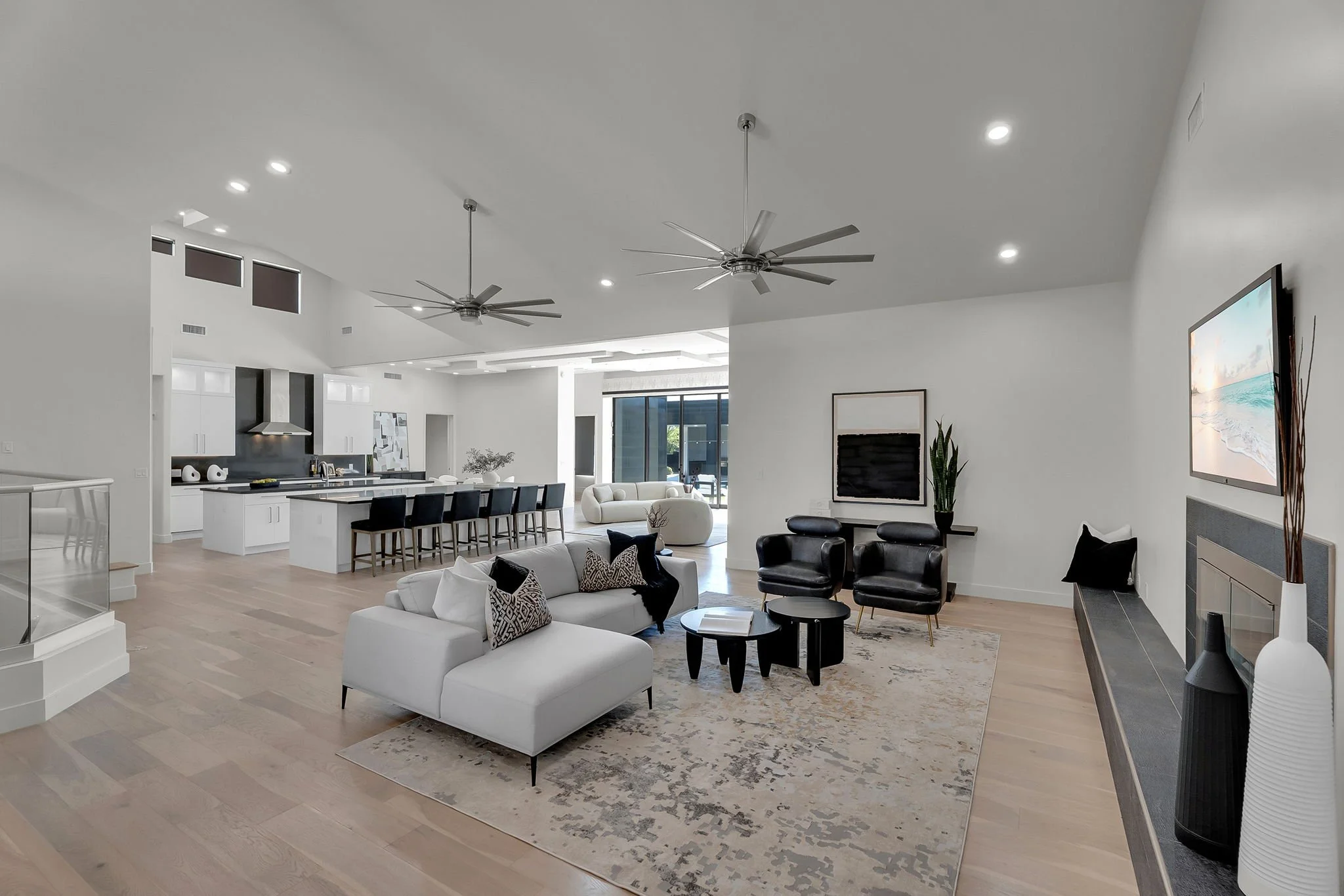 Modern, open-concept living room and kitchen with white walls, light wood flooring, and minimalist furniture. The living room has a white sofa, black armchairs, and a large wall-mounted TV, with decorative vases and a vintage-style rug. The kitchen f