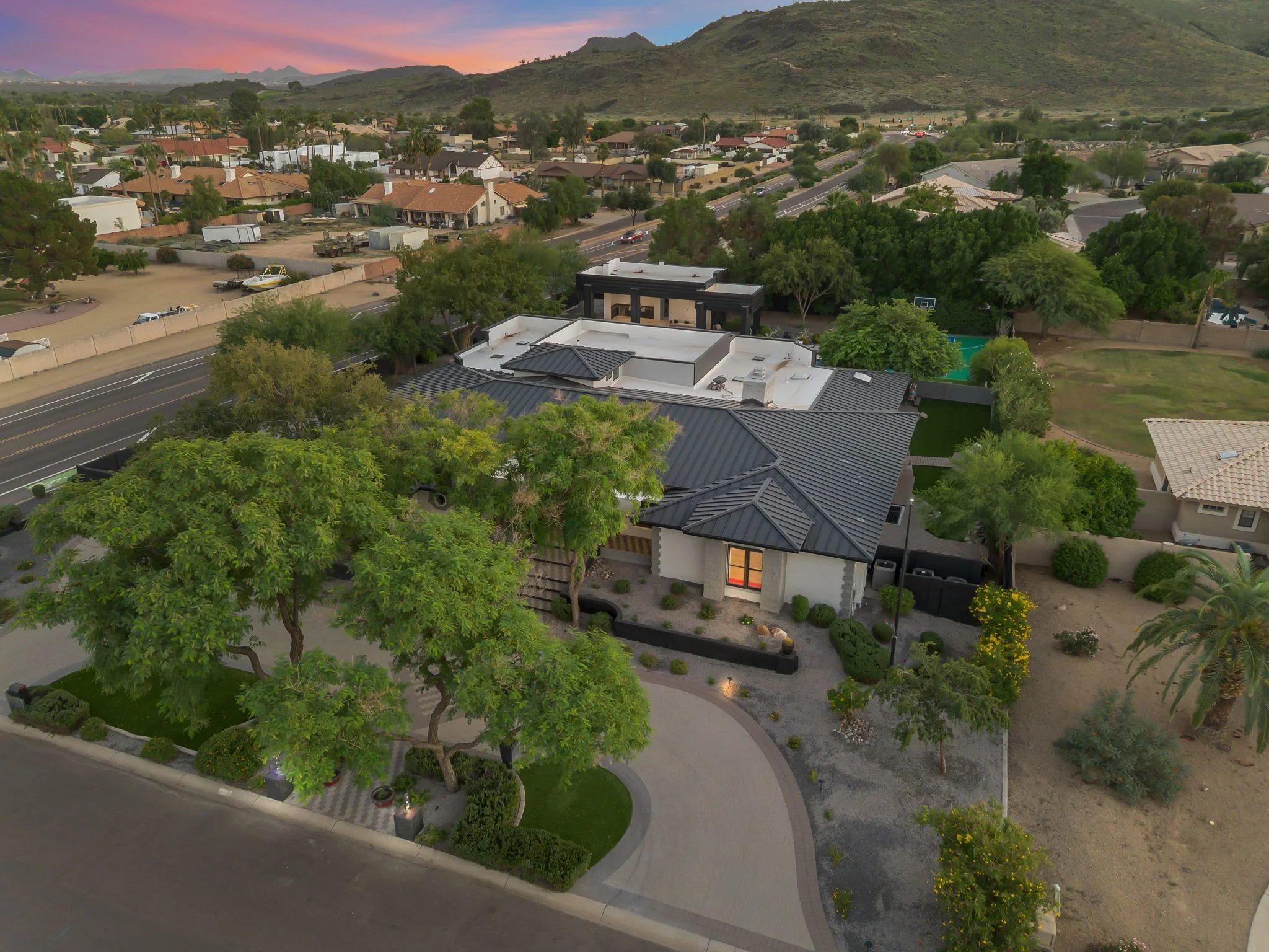 Aerial view of a modern house with a black metal roof, landscaped front yard with trees and bushes, surrounded by neighboring houses and a street, with hills in the background at sunset.