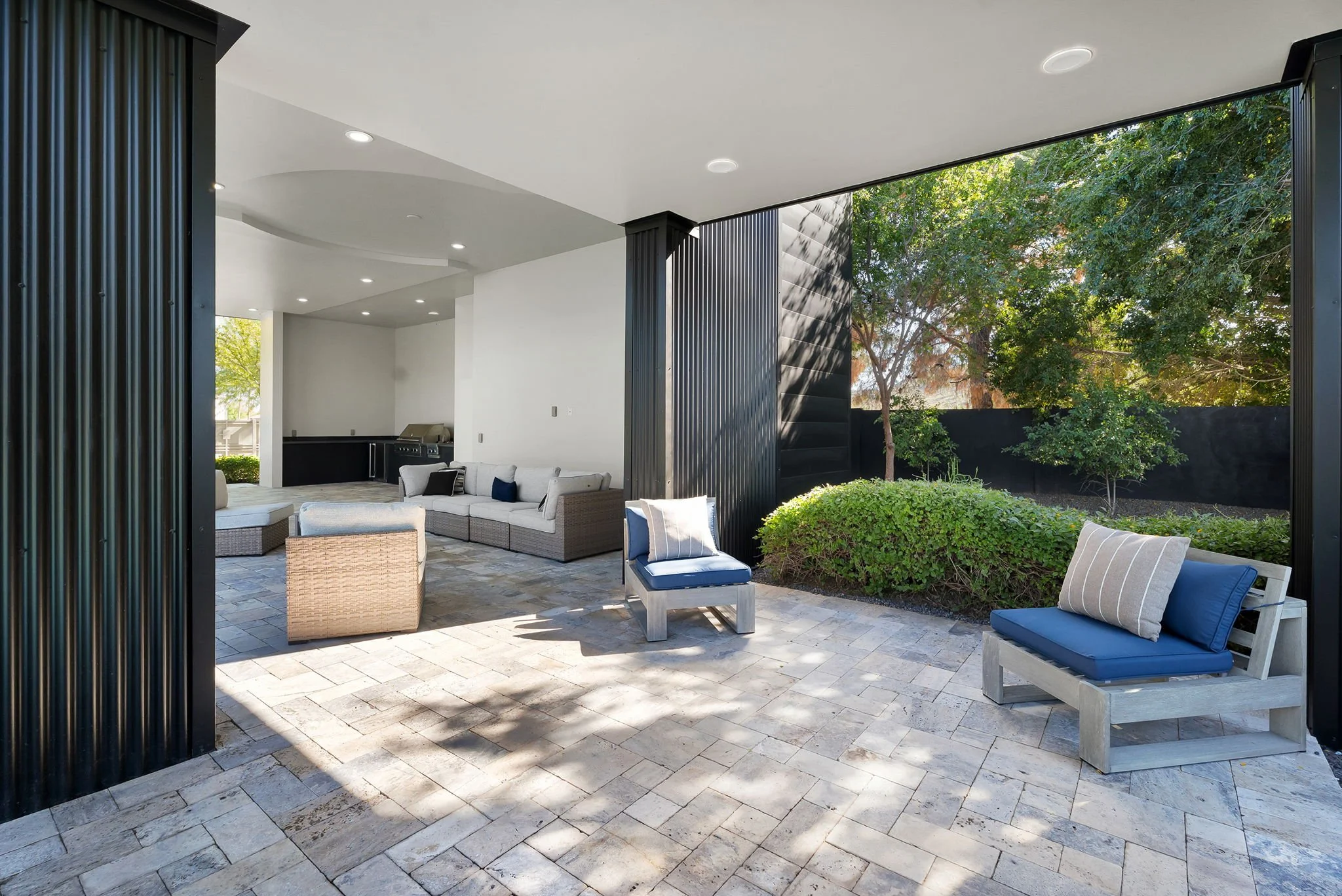 Covered outdoor patio with wicker and wooden furniture, cushions, a built-in grill, and surrounding greenery.