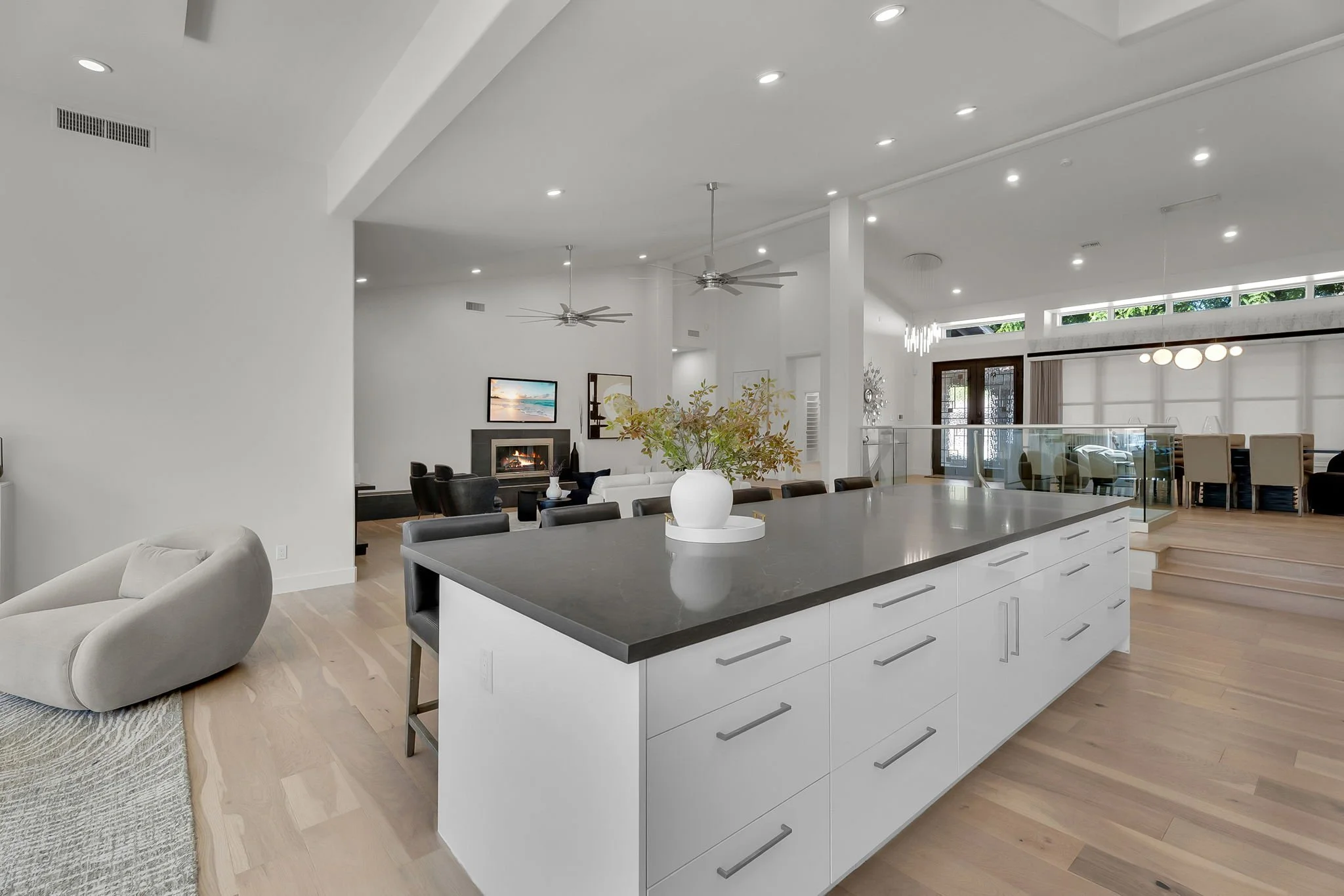Open-concept living space with a kitchen island, white cabinetry, gray countertop, and a large vase with greenery. In the background, there is a living room with a fireplace, television, and seating area, all decorated in modern style with light wood