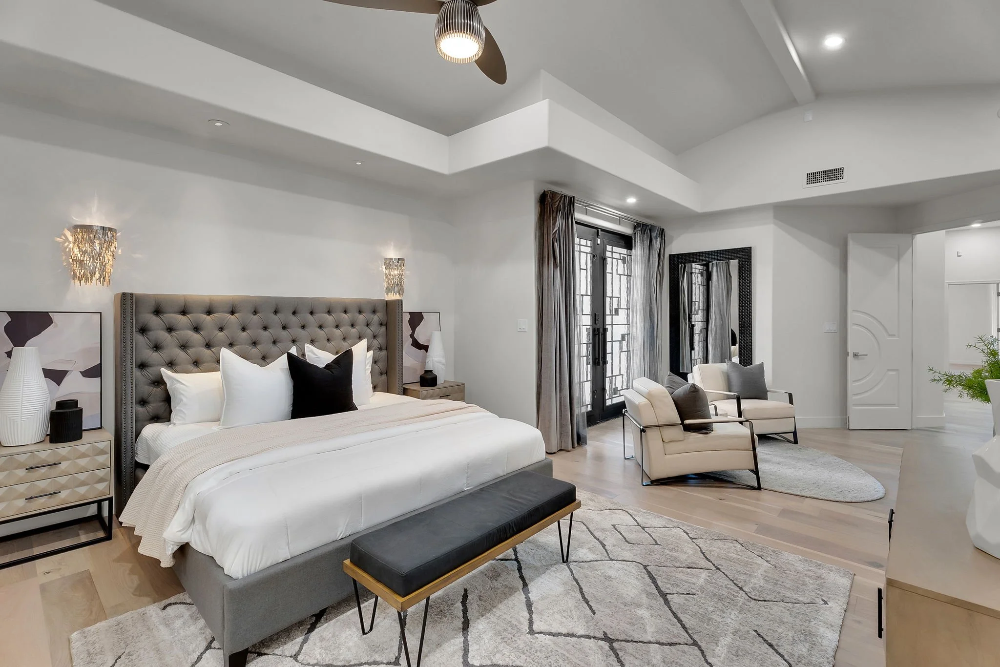 Modern bedroom with a large bed, tufted headboard, nightstands, armchairs with pillows, full-length mirror, curtains, and wooden flooring.