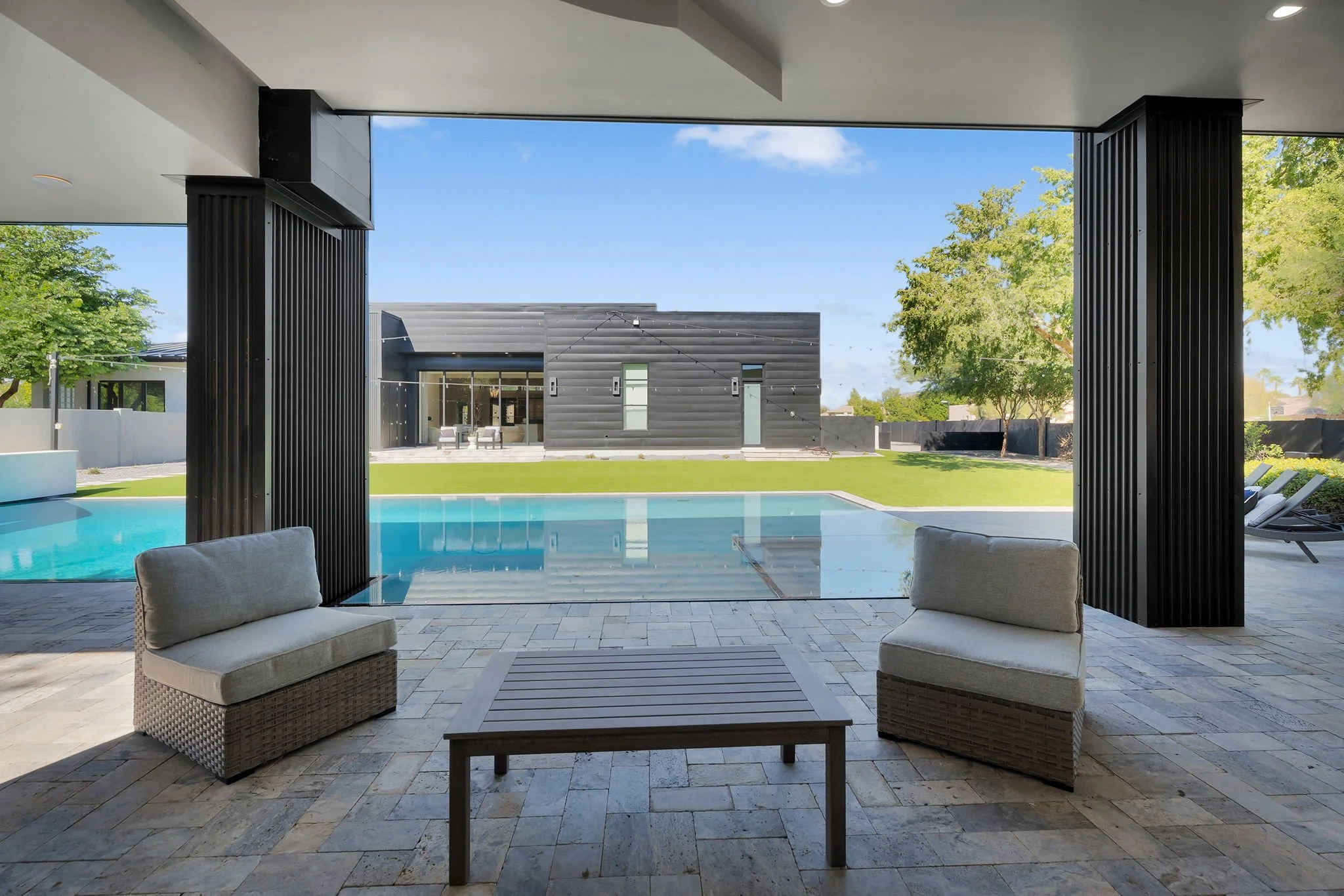 Modern outdoor patio with seating area overlooking a backyard with a swimming pool, green lawn, trees, and a contemporary black building in the background under a blue sky.