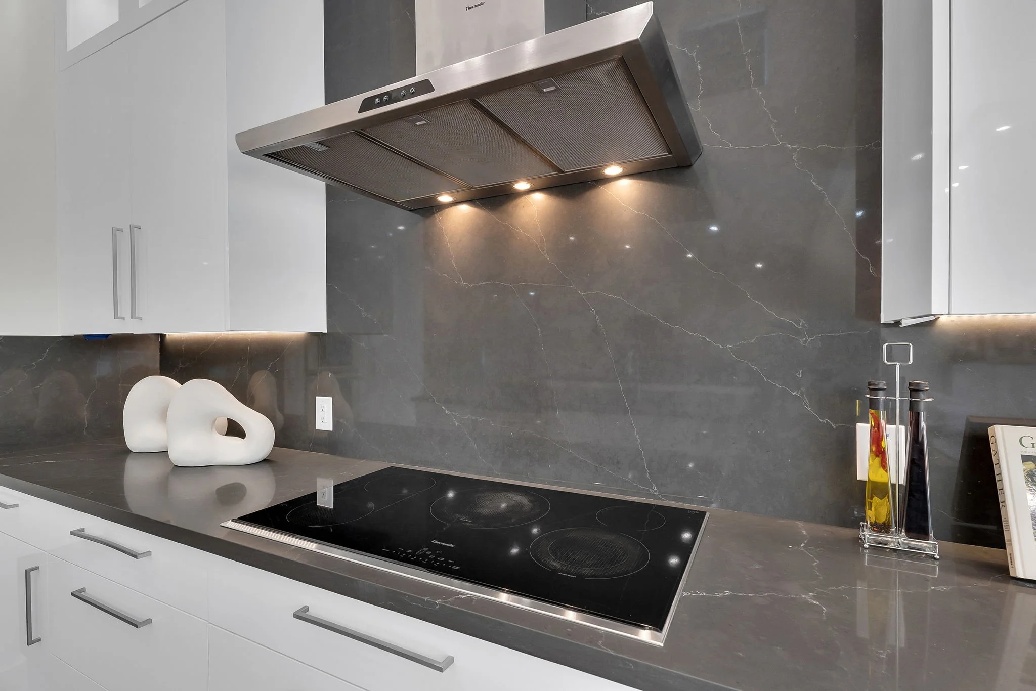 Modern kitchen with gray marble backsplash, white cabinets, and a black induction cooktop. Features decorative white ceramic objects, a paper towel holder, and oil bottles.