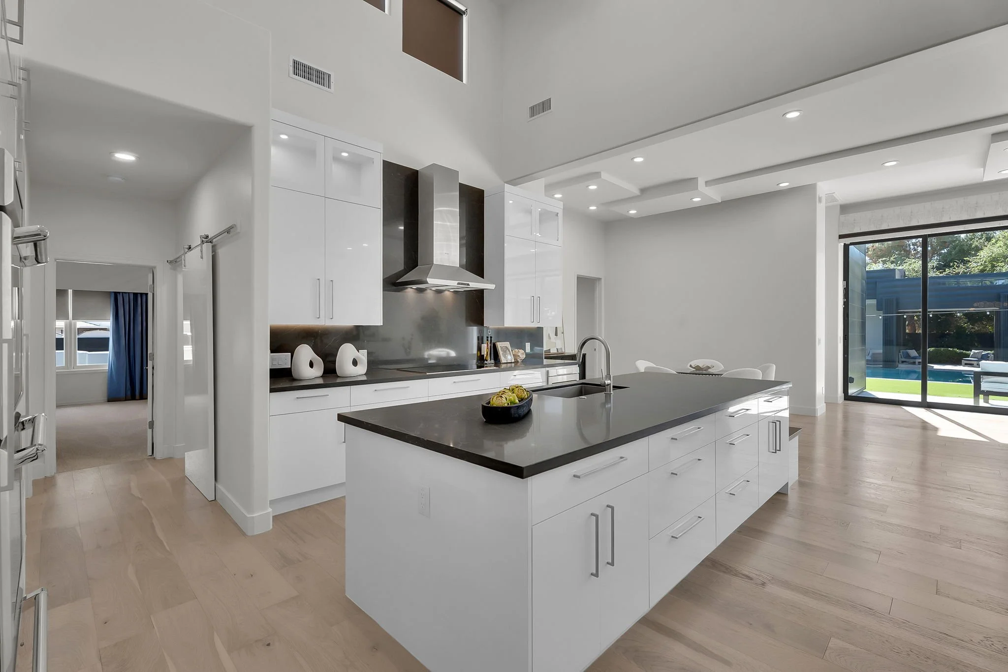 Modern kitchen with white cabinets, a large island with a black countertop, stainless steel appliances, and sliding glass doors leading outside, featuring light wood floors and recessed lighting.