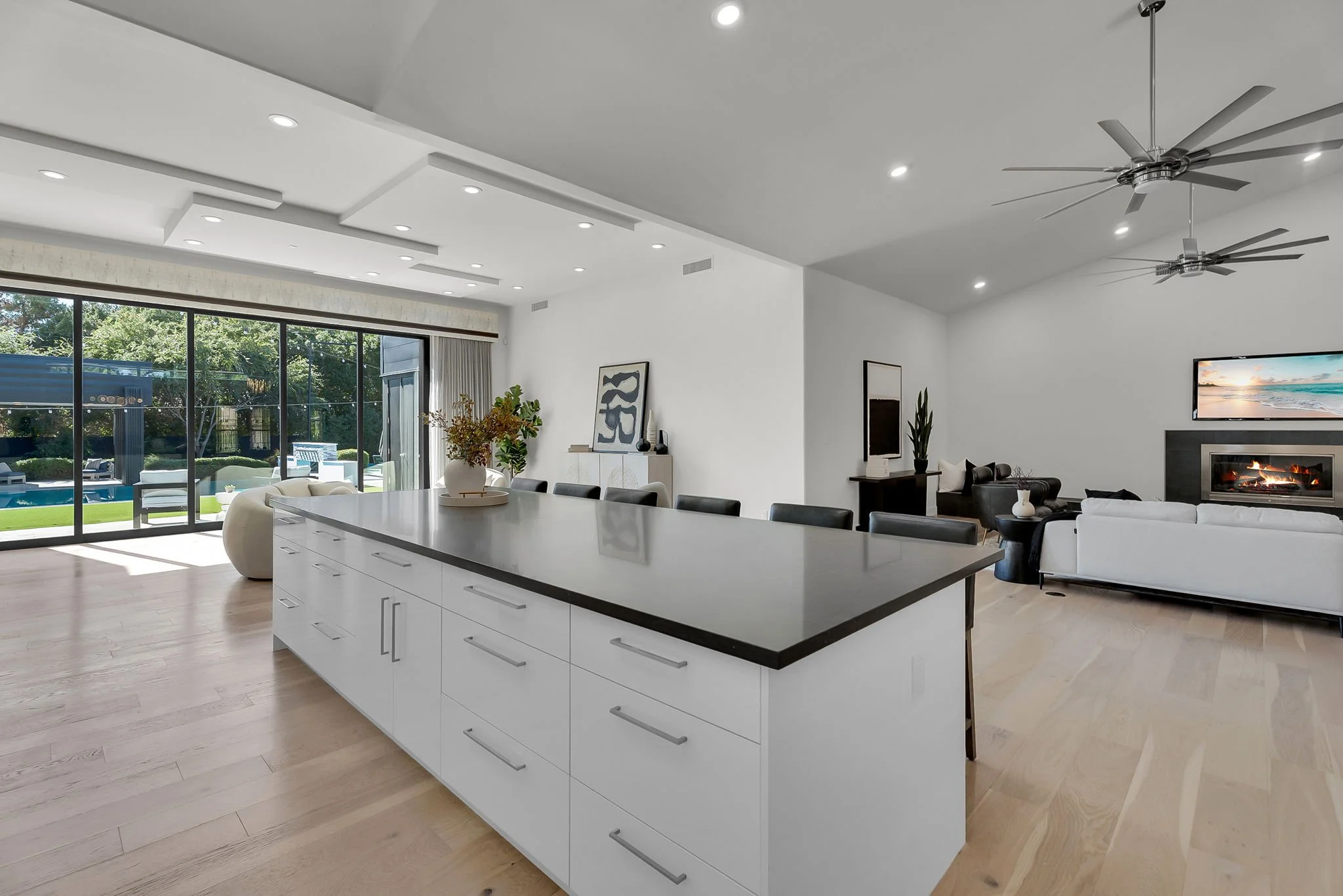 Modern open-plan living room and kitchen with large sliding glass doors, light hardwood floors, white walls, ceiling fans, a fireplace, a wall-mounted TV, and contemporary furniture.