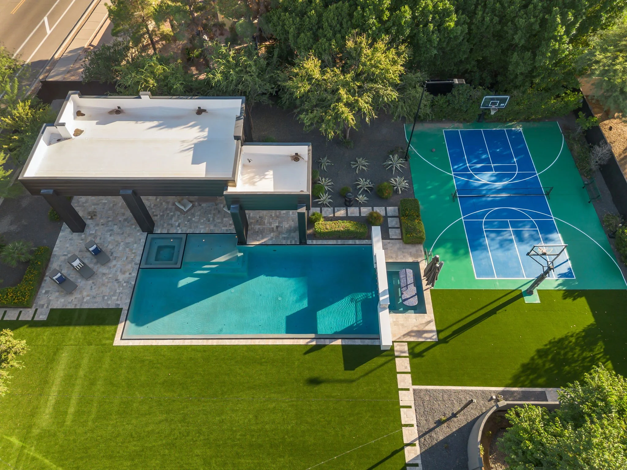Aerial view of backyard with swimming pool, outdoor seating area, two-story building, and a basketball court surrounded by trees and greenery.