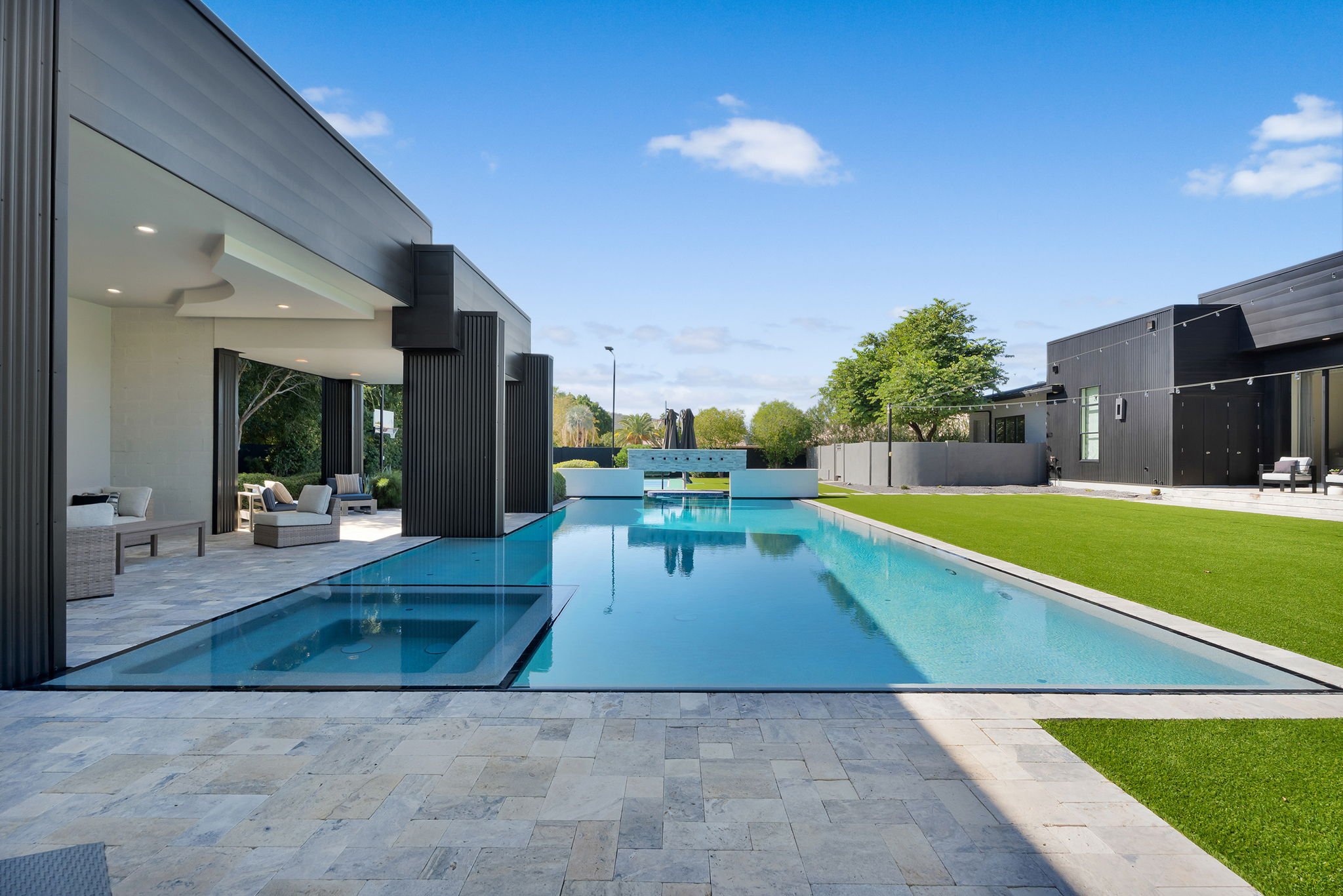 Modern house with an infinity pool, patio seating area, and well-maintained lawn under a bright blue sky.