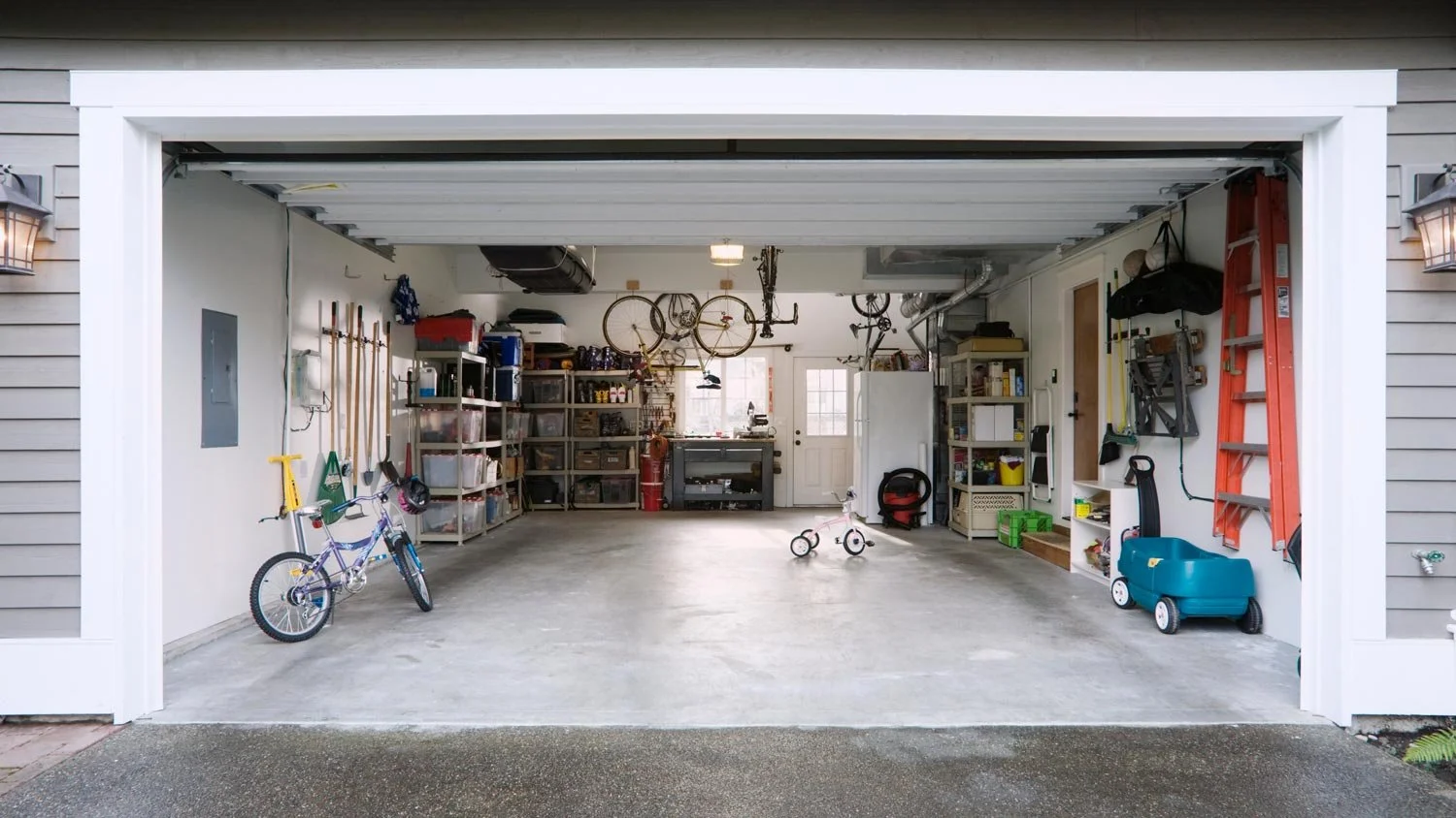 Clean garage with storage shelves, children's bicycle and toys, tools, ladder, and bicycles hanging from ceiling, illuminated by natural light from windows and door.