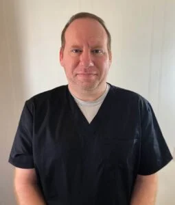 A man dressed in black scrubs standing against a plain light-colored background, smiling slightly.