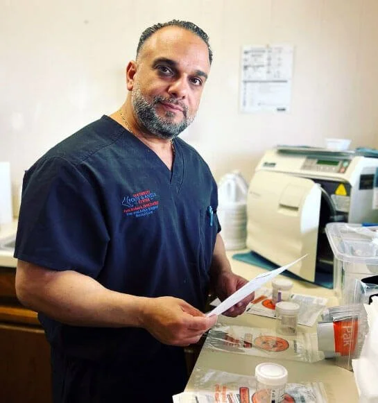 A male medical professional in navy scrubs standing at a medical countertop with lab supplies and a lab machine, holding a piece of paper.