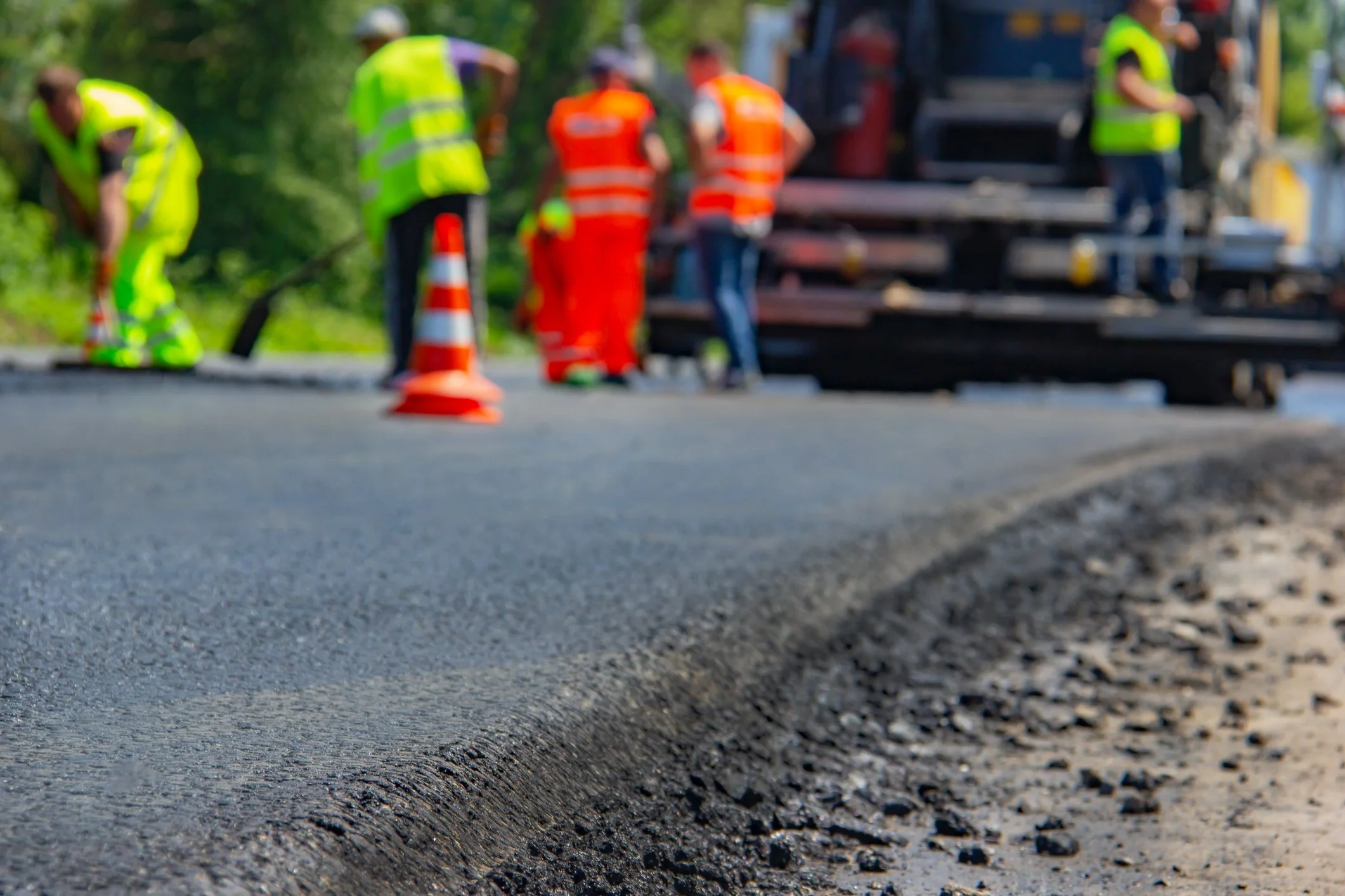 Road construction workers in safety vests and helmets working on a paved road, with orange traffic cones and construction equipment nearby.