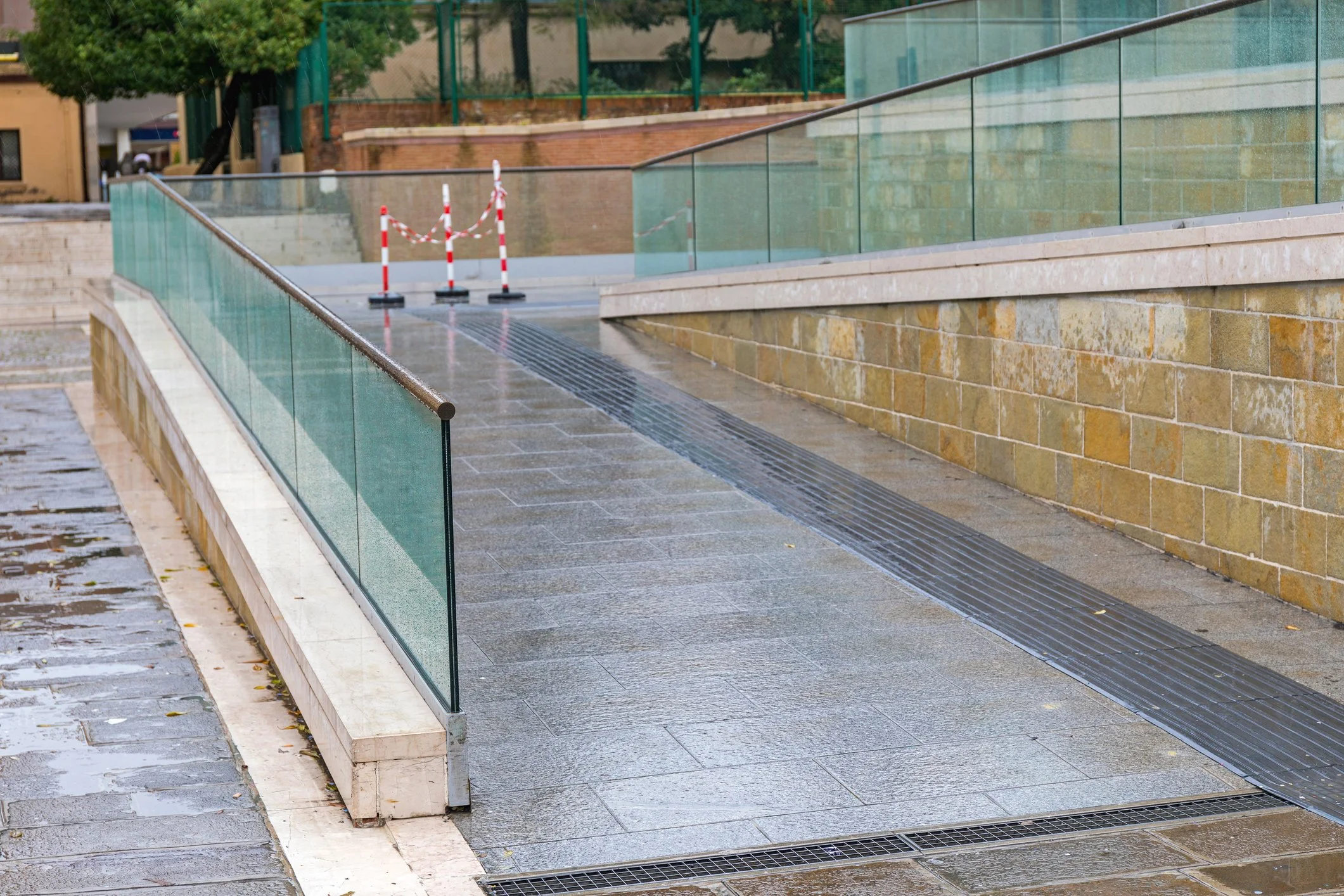 Rain-soaked urban sidewalk with a glass railing, a stone wall, and a closed area marked by red and white barricades.