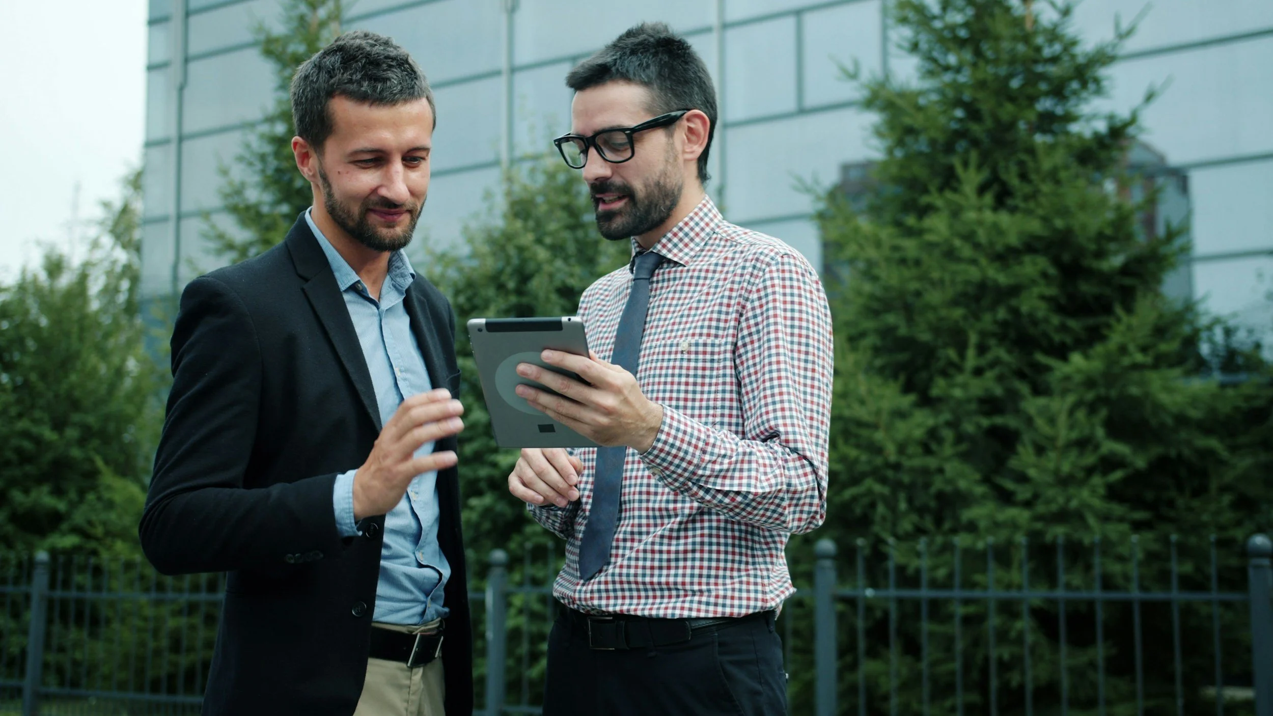 Two men in business casual attire standing outdoors in front of green trees and a building, looking at a handheld device together.