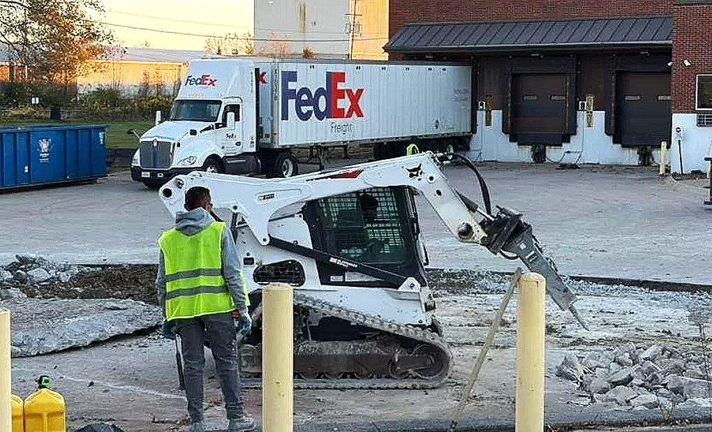 Construction worker in a yellow safety vest operating a small skid steer loader on a damaged asphalt surface outside a building, with a FedEx truck parked in the background.