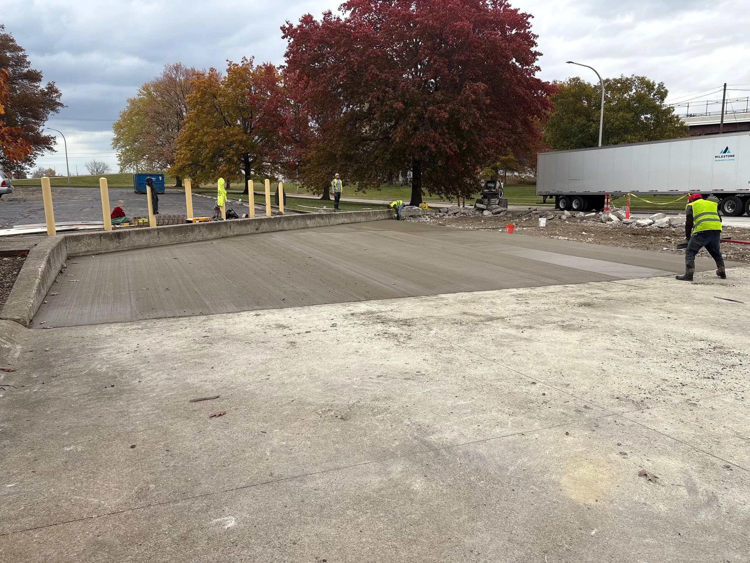Construction workers paving a new section of a road or parking lot, with trees with red and orange leaves in the background and a large truck parked nearby.