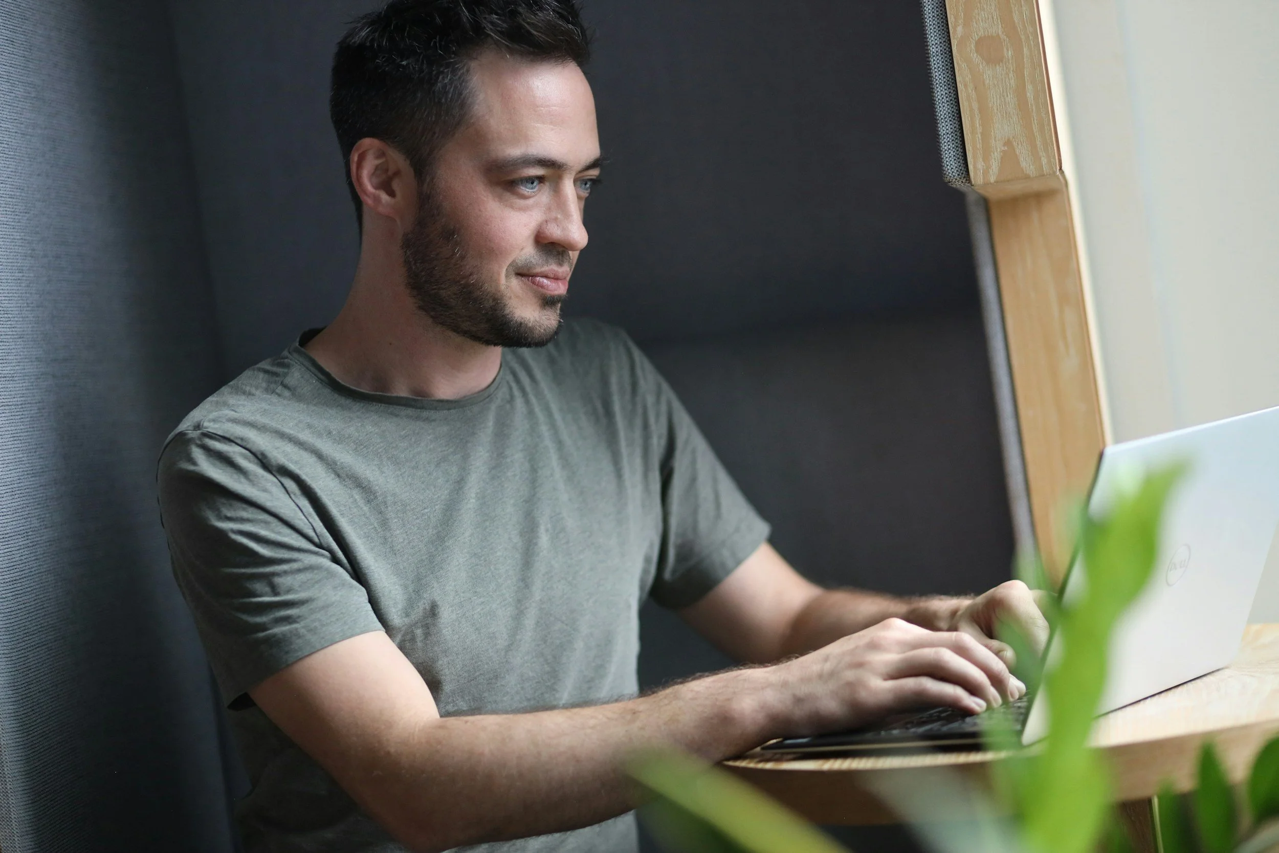 A man with short dark hair and a beard sitting by a window, working on a laptop with a green plant in the foreground.