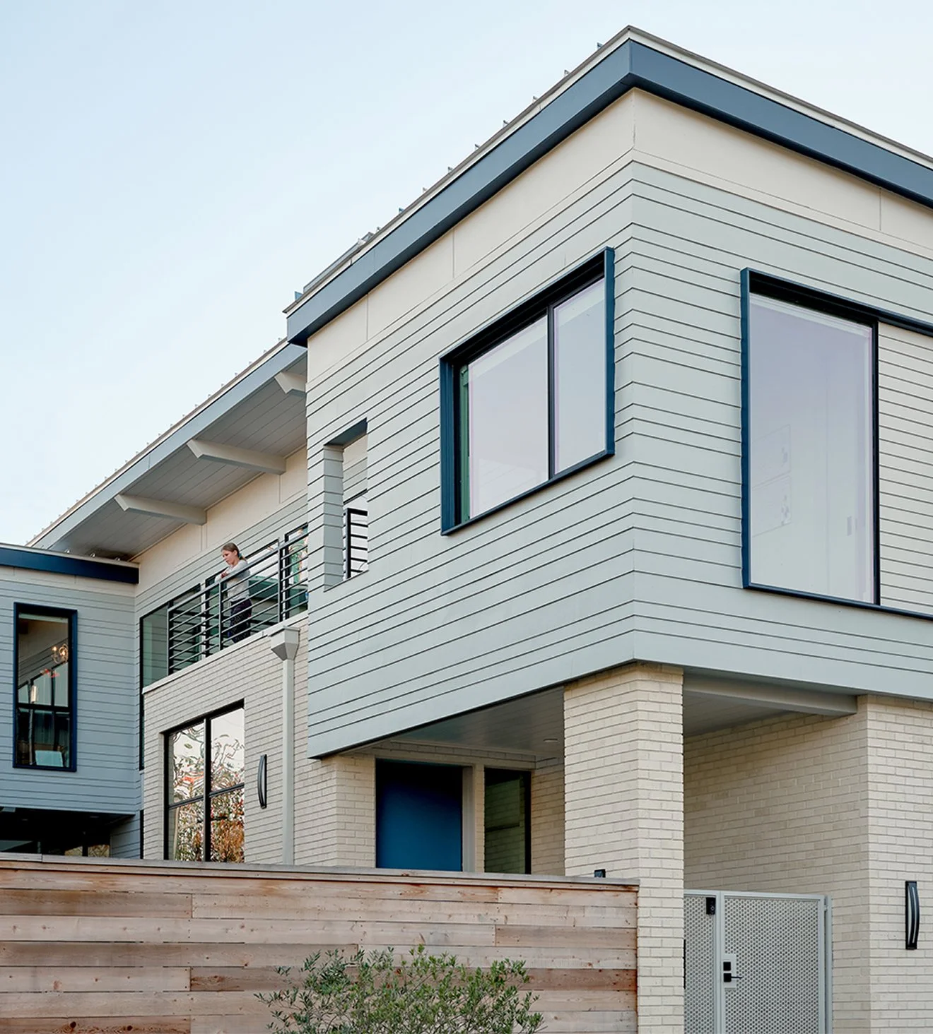 Modern multi-story residential building with large windows, a balcony, and a wooden fence in the foreground.