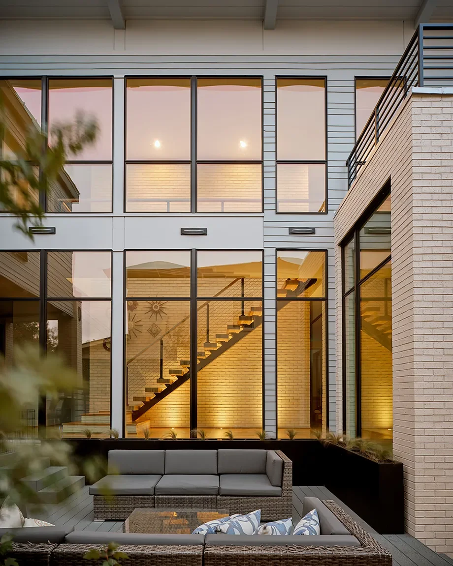 Modern multi-story house with large glass windows, showing an interior staircase, lit warmly from inside, with a cozy outdoor seating area featuring a couch and chairs with pillows.